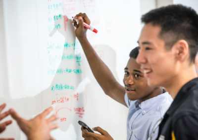 Three young men, students, work on math problems on a communal whiteboard.