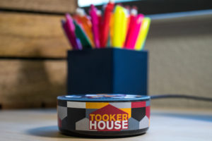 A Tooker House-branded Echo Dot (TM) sits on a desk in front of a box of pencils and highlighters.