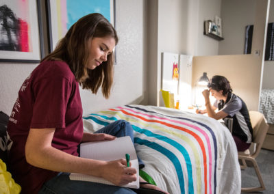 Two young women are studying in their room. One sits at her desk and one on her bed.