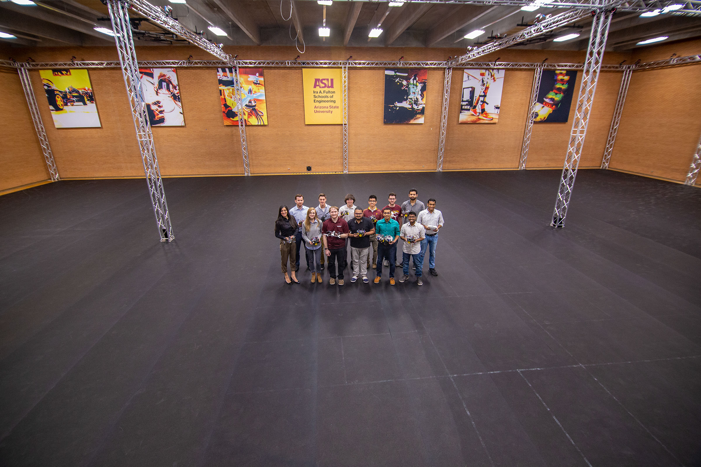 A group of about 20 researchers stand in the middle of the immense ASU Drone Studio, surrounded by tons of open space.