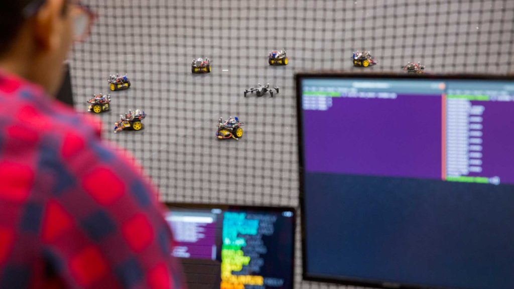 A researcher sits at a computer screen behind a net separating him from the testing area. He is controlling a robot swarm with the computer.
