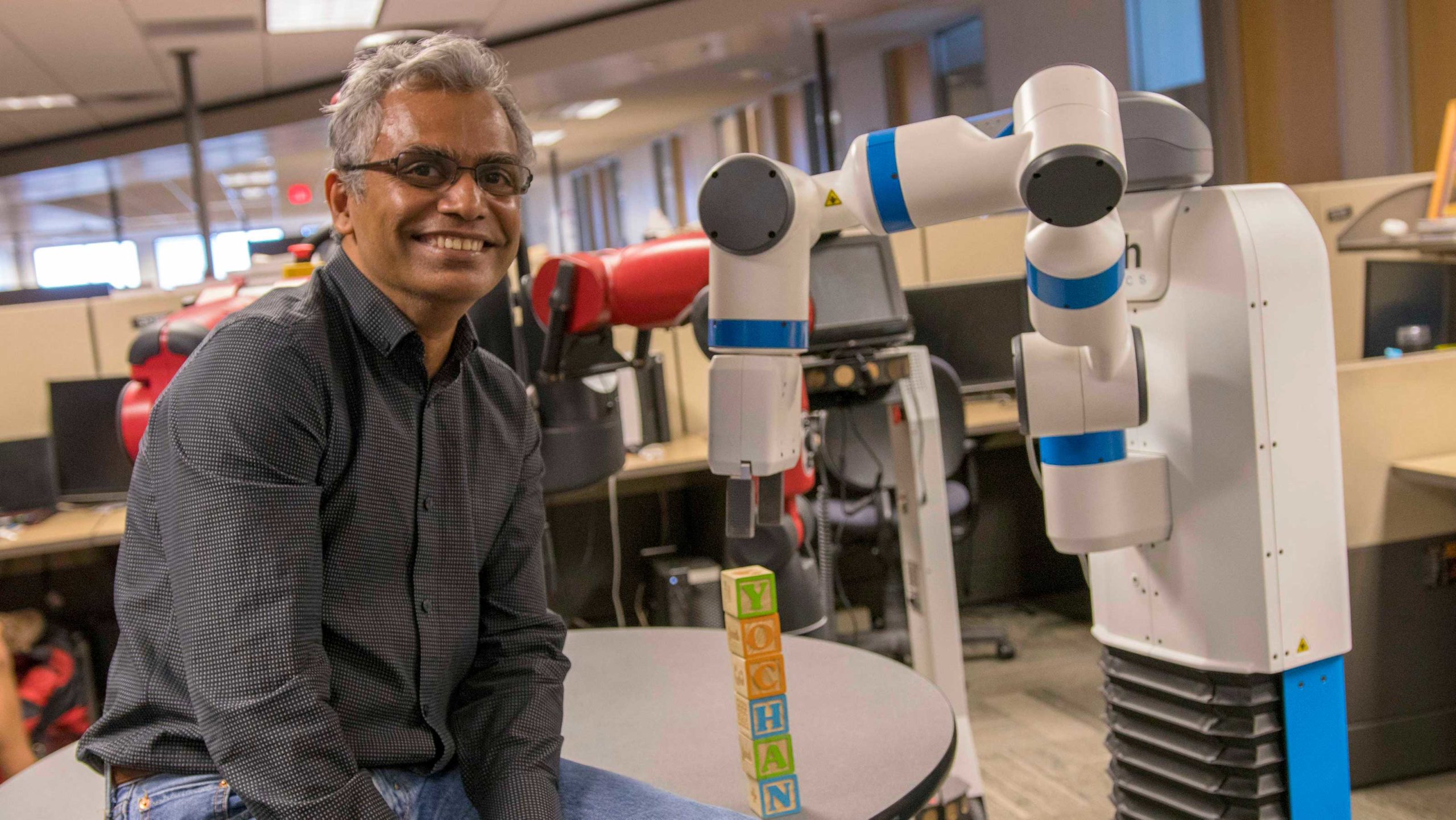 Rao Kambhampati poses for the camera in his lab next to a robot that has just stacked some alphabet blocks