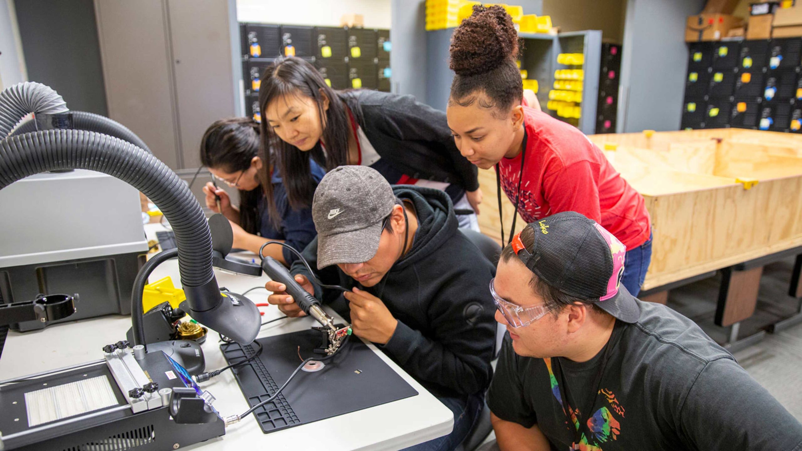 Five undergraduates work together soldering a small robot component at the ASU 2019 Robo Hackathon
