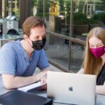 Two students wearing masks while sitting outside and sharing a laptop.