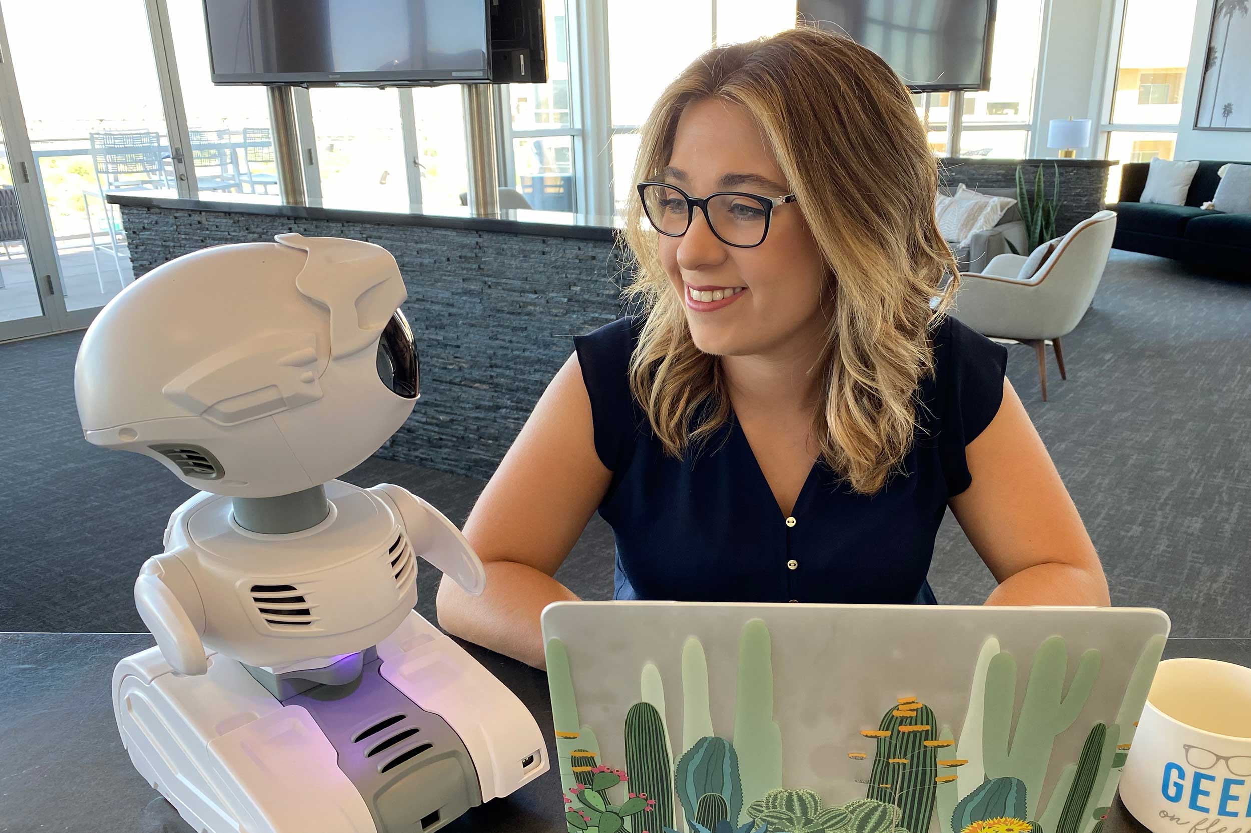 Female PhD student student Jordan Miller works at a laptop, smiling towards a small white robot sitting next to her.