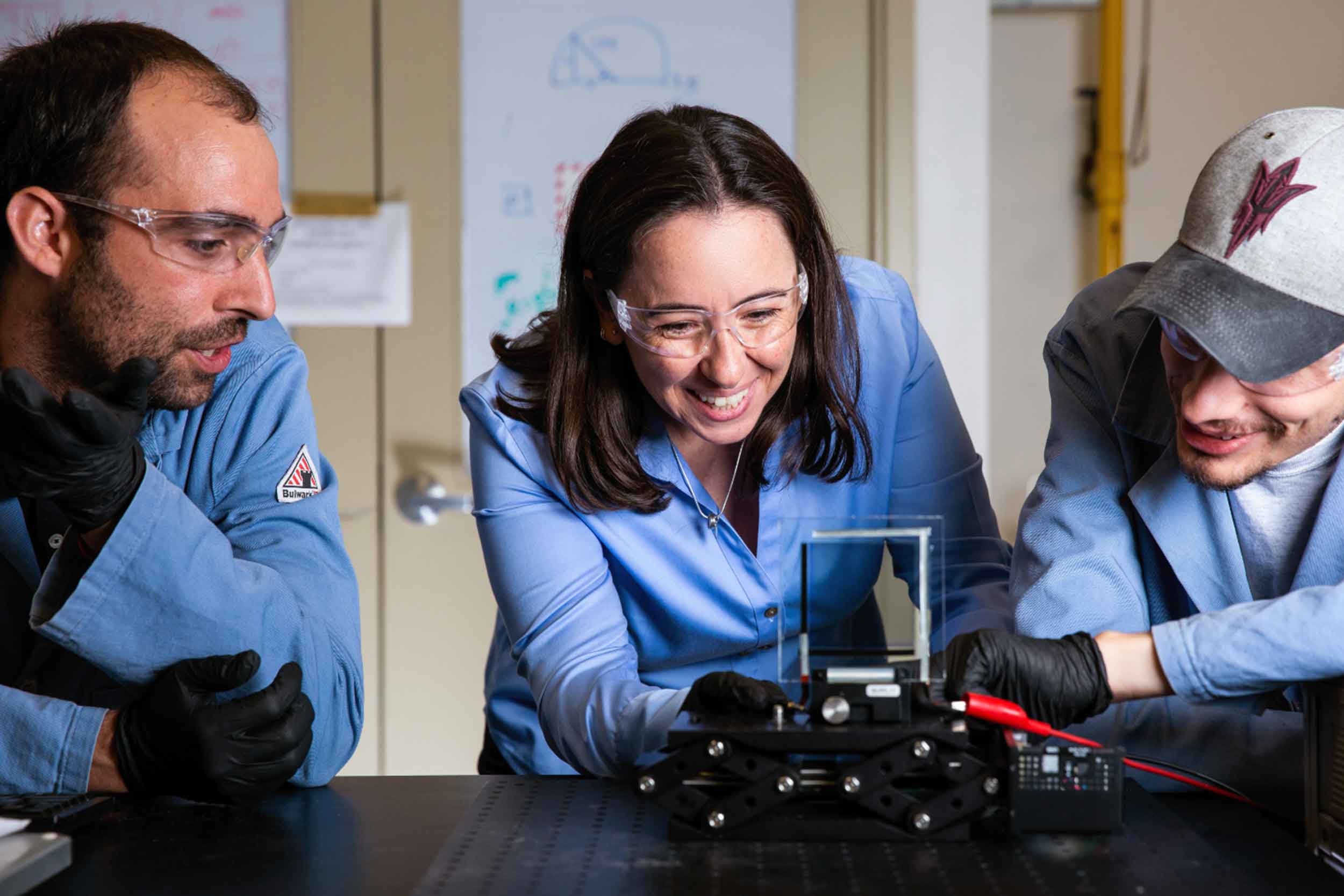 Mariana Bertoni and two researchers lean in at the lab bench looking at a device they are working on