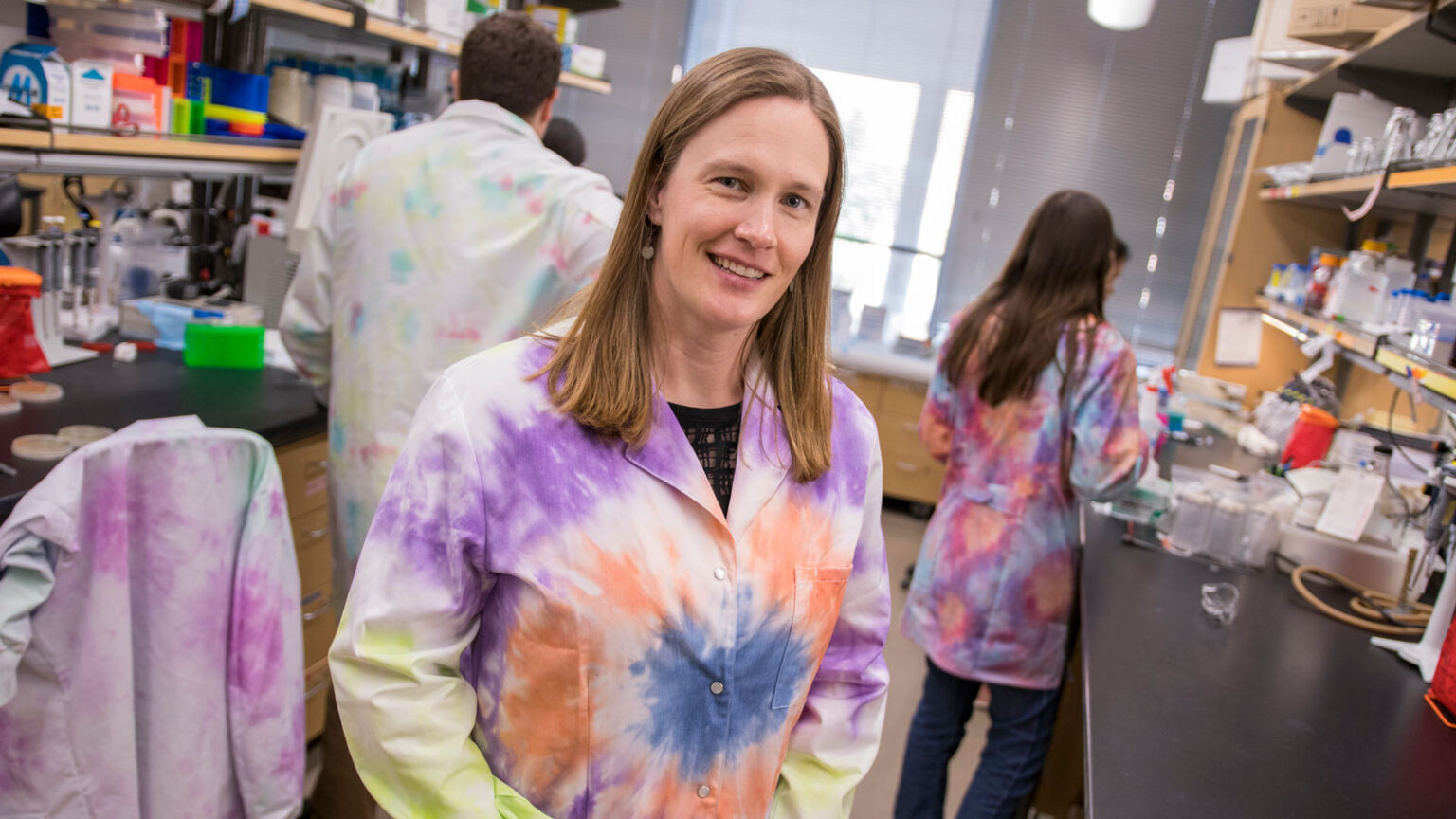 Sarah Stabenfeldt, wearing a tie-dye lab coat, stands in her lab with other researchers