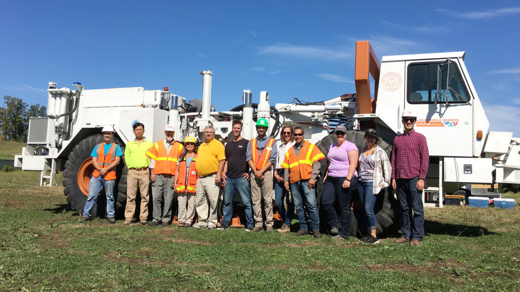 A dozen people stand side by side, some in construction vests, in front of the huge T-Rex ground shaker system