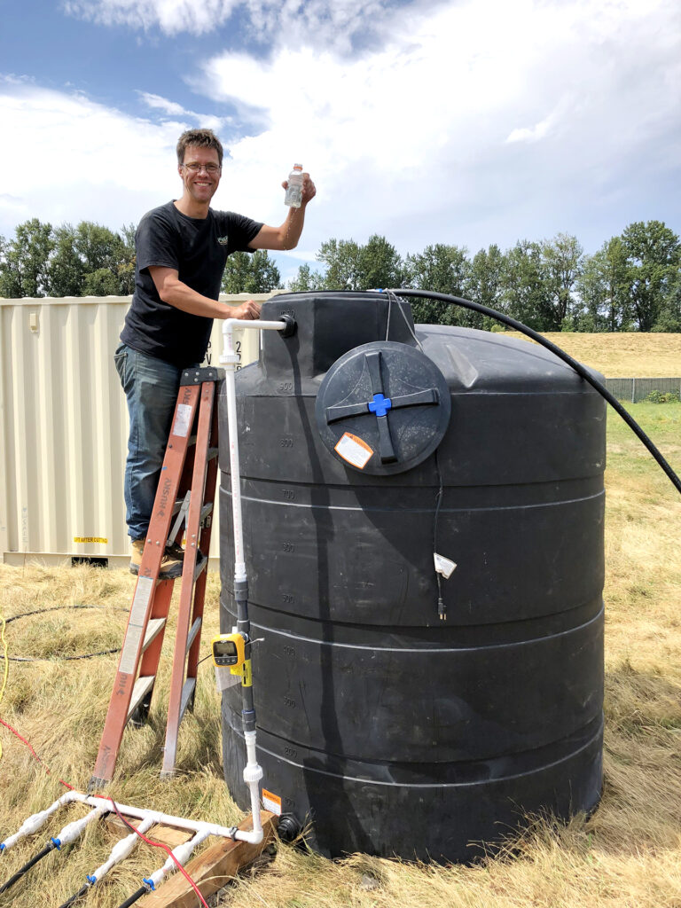 Leon van Passen stands on a ladder outdoors leaning against a large, tall black plastic water storage tank holding a water sample in a bottle