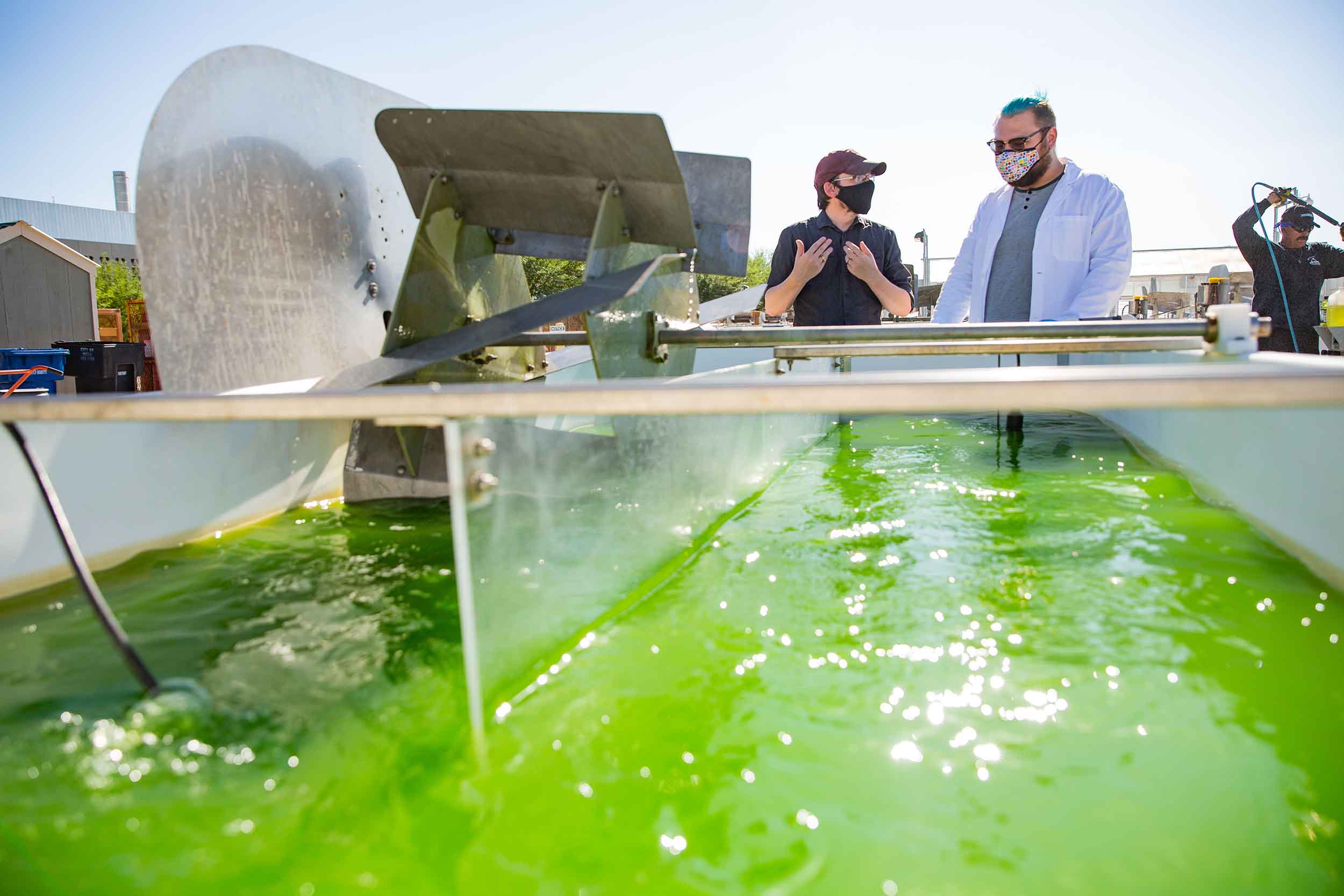 Taylor Weiss and Duane Barbano stand outside in the bright Arizona sun next to a gigantic vat of churning water full of bright green algae