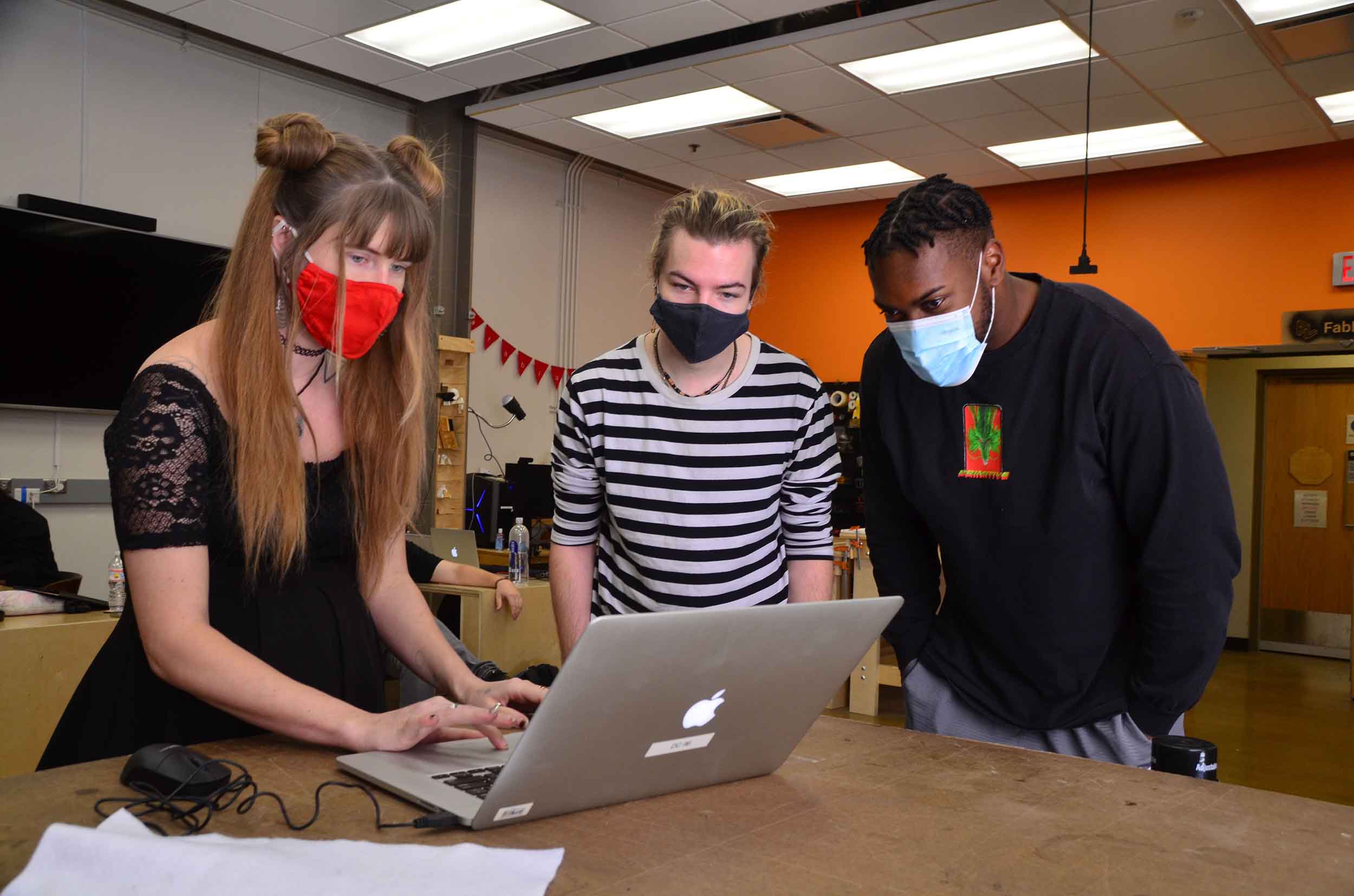 Three students in masks stand looking at a laptop during a FabLab workshop.