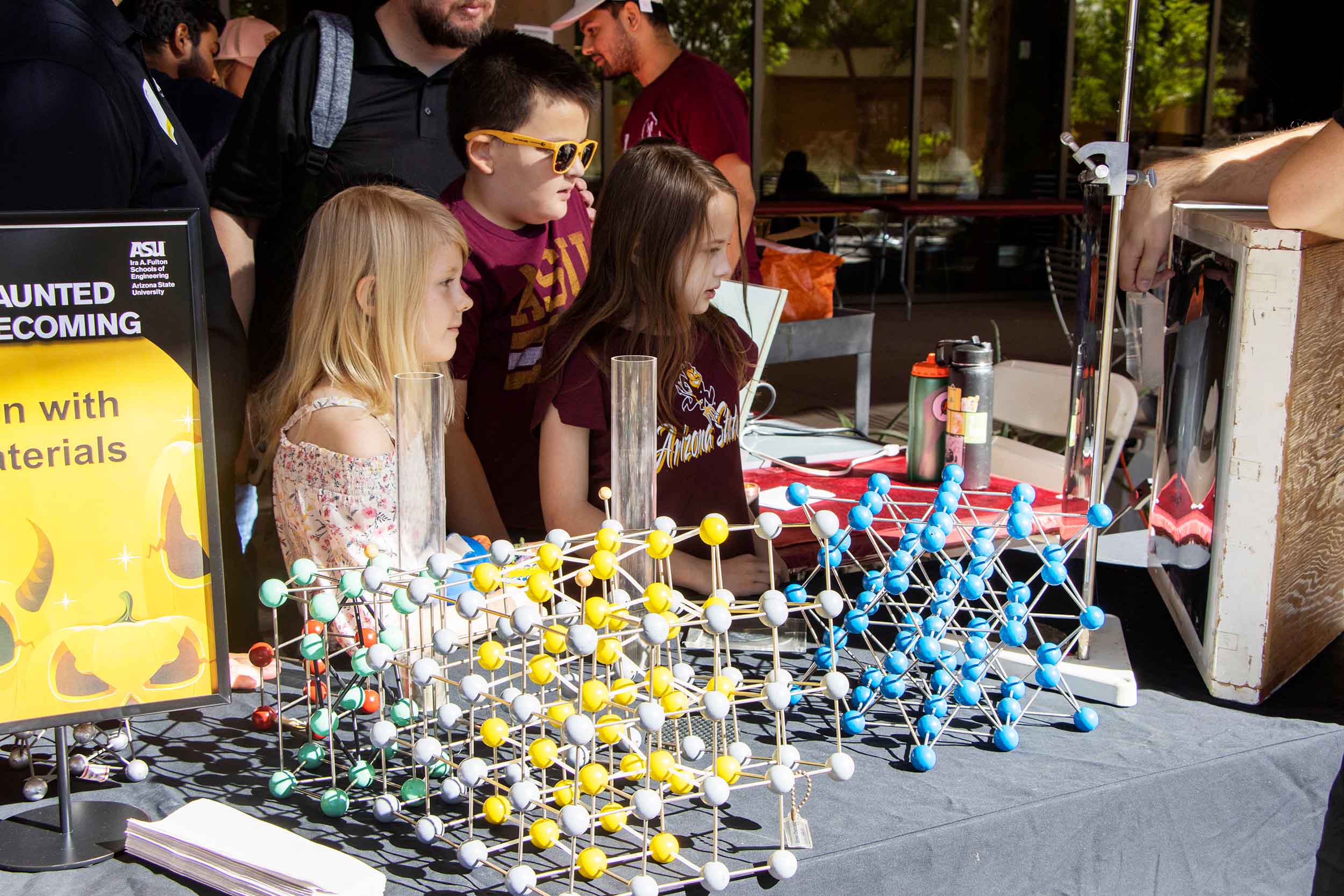Three children with their families stop to learn a little bit about materials science, surrounded by molecular models and graduated cylinders and a mystery in a box demonstration by members of the Material Advantage student org.