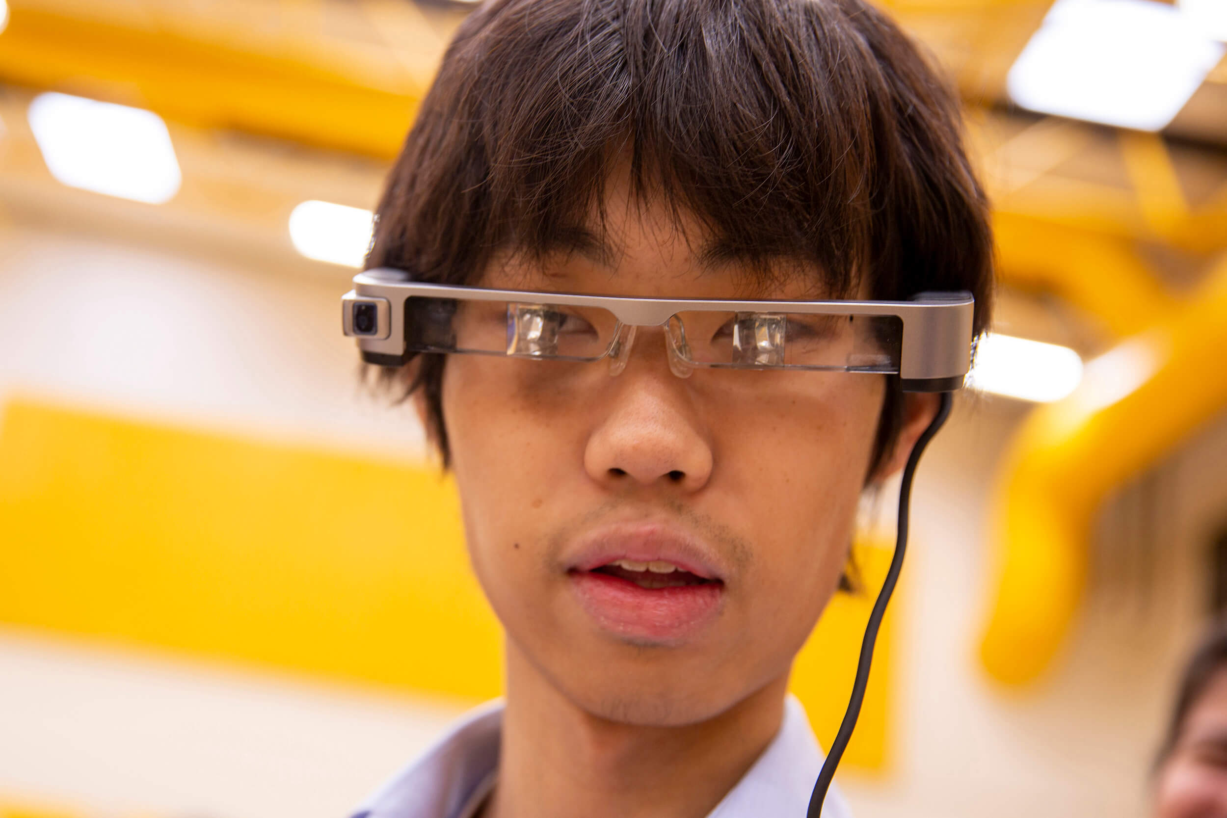 Close up of an undergraduate student at a student capstone (final project) event wearing a futuristic, thin type of electronic eyewear with small square lenses attached to a cable, which were part of his research presentation "Dynamic Signal Model for Immersive Therapeutic Biofeedback"