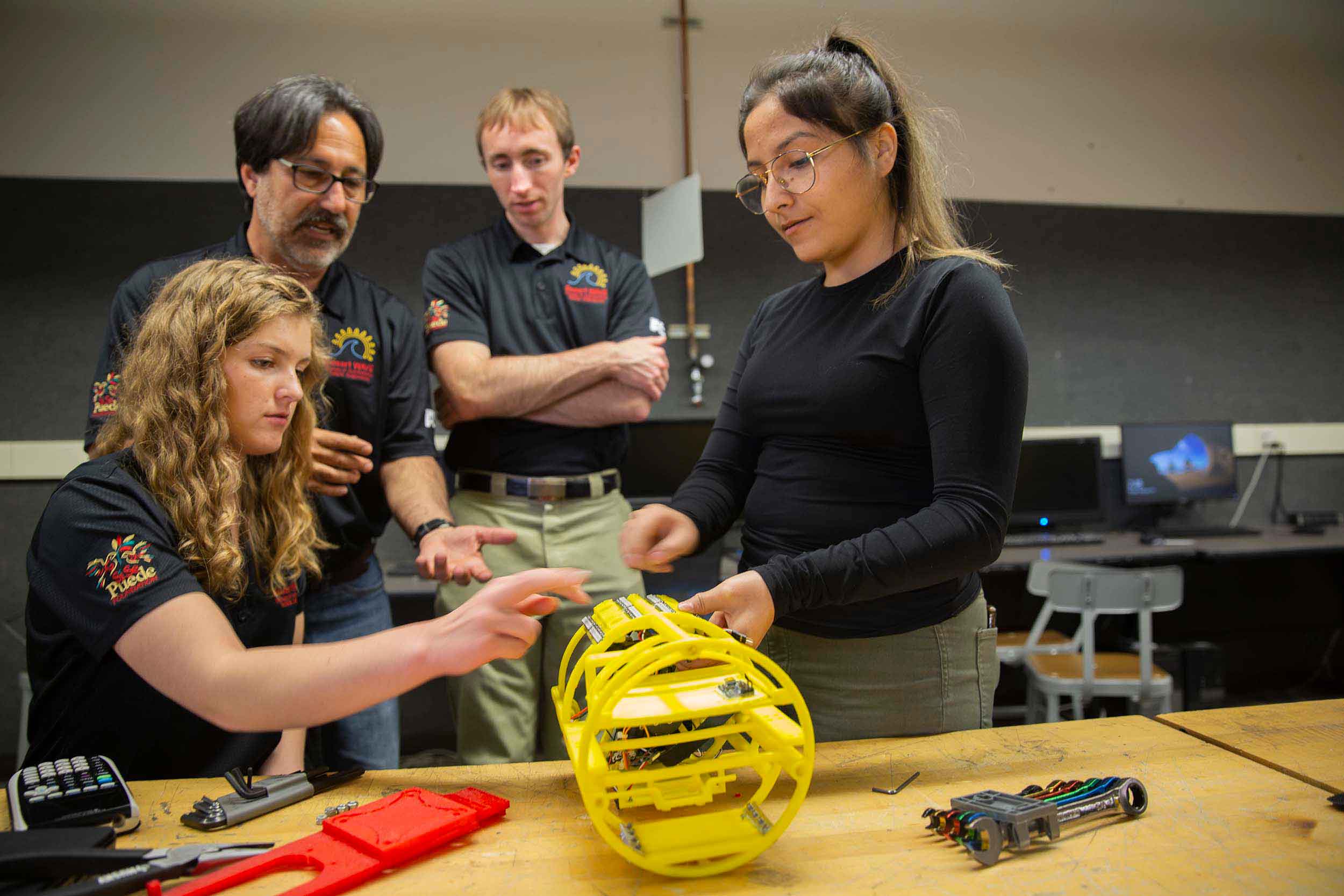 Two undergraduate members of the all-female Desert WAVE underwater robotics team work on their robot in a workshop with two mentors.