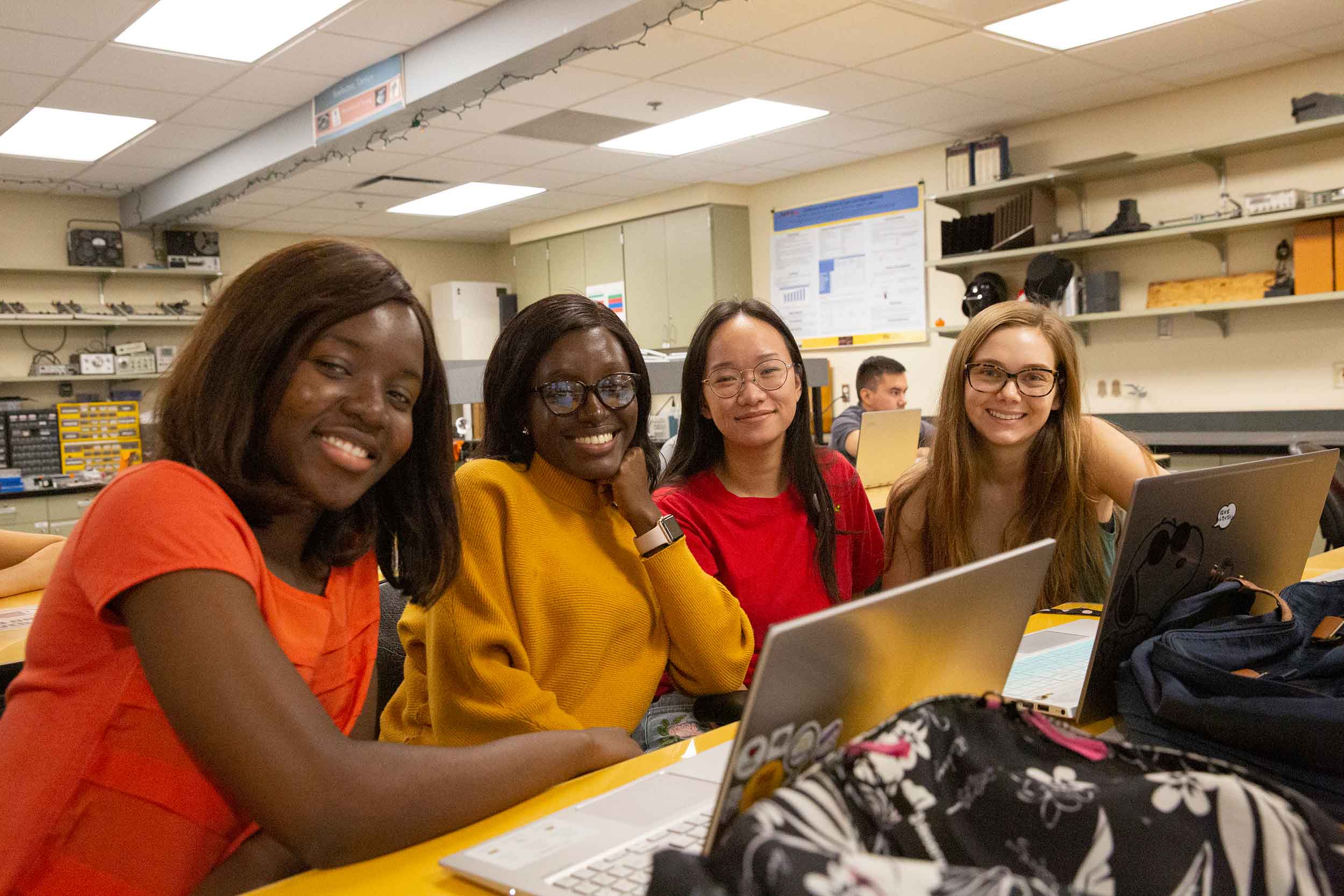 Four undergraduate women take a break from their research to smile for the camera while sitting at a long lab table with their laptops.