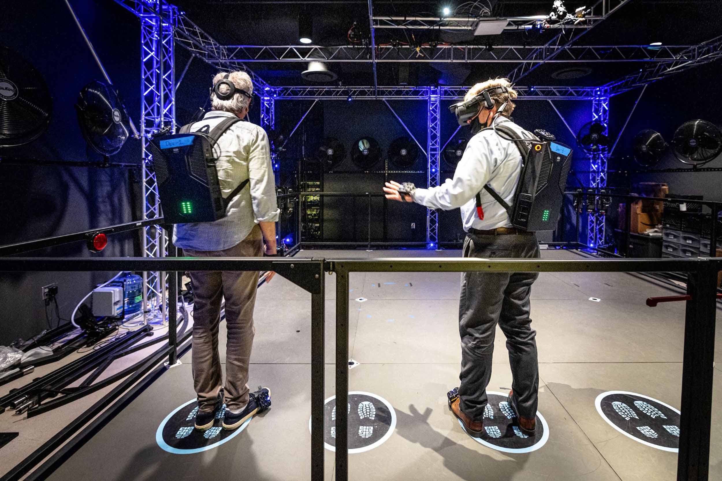 Walter Parkes and Michael Crow stand side by side in an ASU Dreamscape pod wearing all the VR equipment needed to experience the Theta Labs project (VR goggles, backpack, hand and foot equipment)