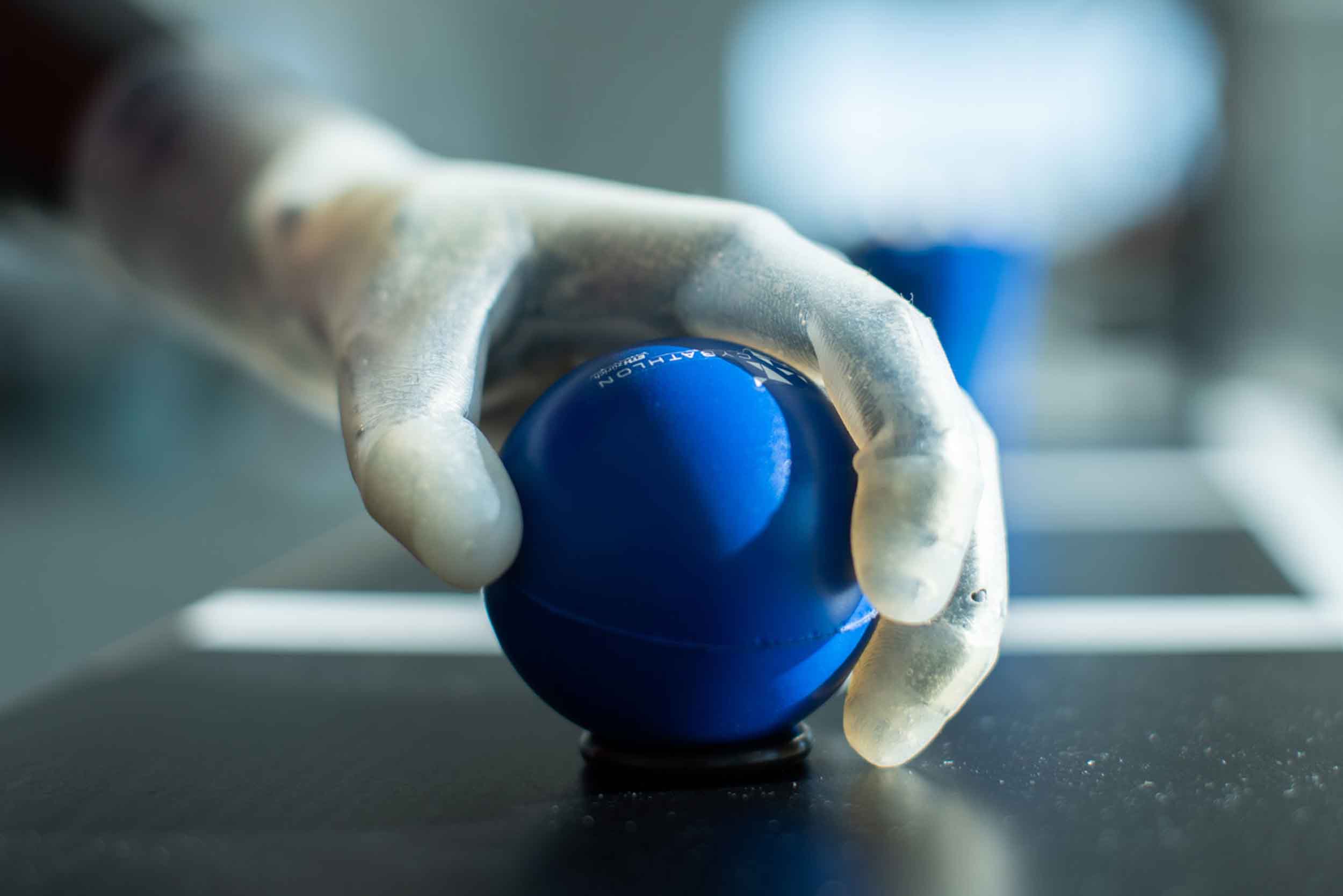 The SoftHand Pro prosthetic hand grips a blue ball. The hand is covered with a clear silicone material and resembles a human hand