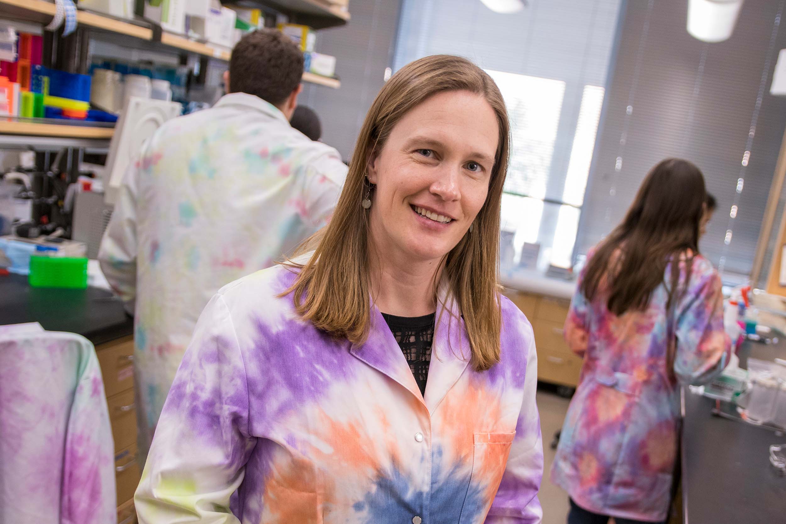 Sarah Stabenfeldt, sporting a colorful tie-dyed lab coat, smiles for the camera in her lab among other researchers