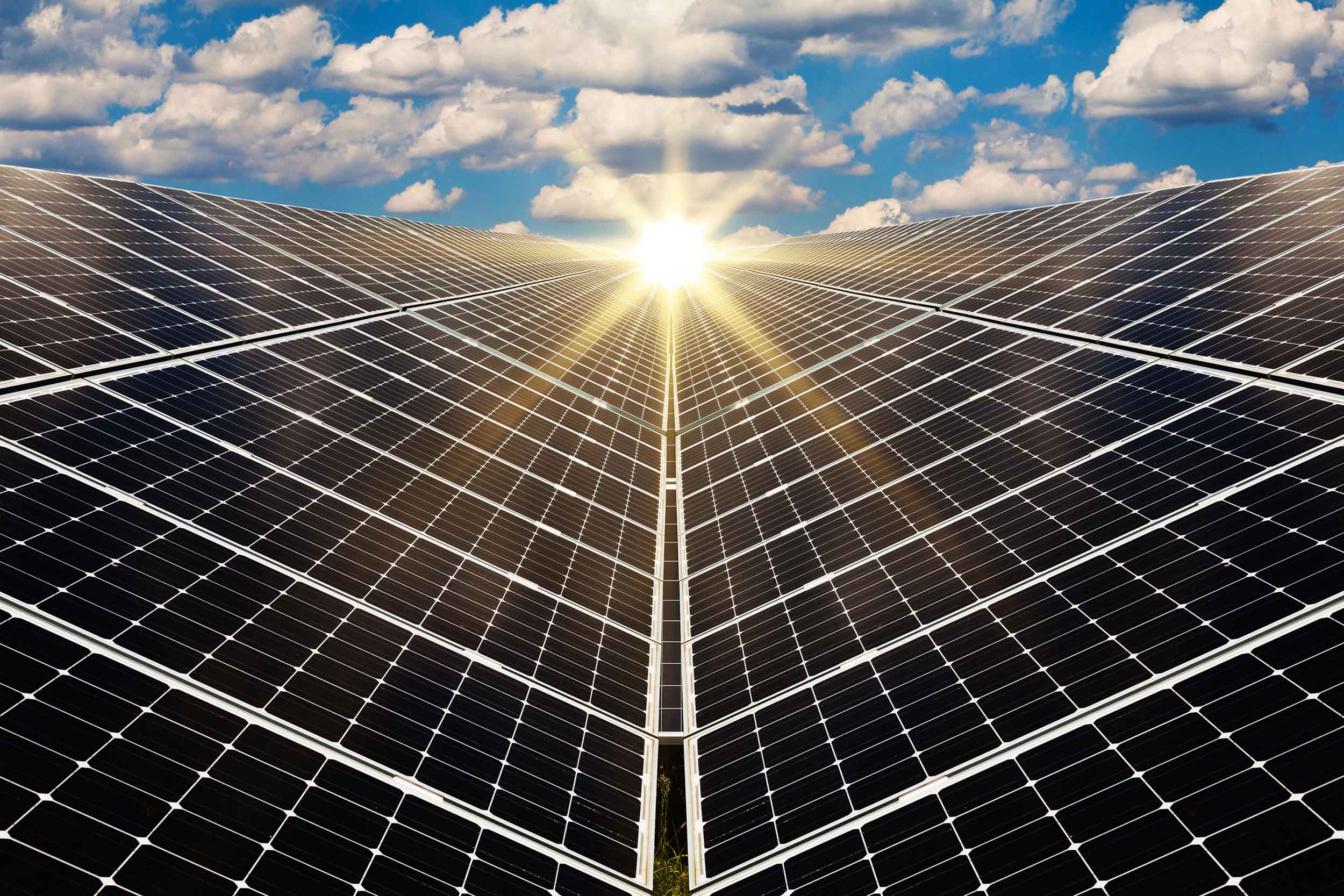 Stock photograph of the sun shining ton a huge array of solar panels with a blue sky background full of puffy clouds