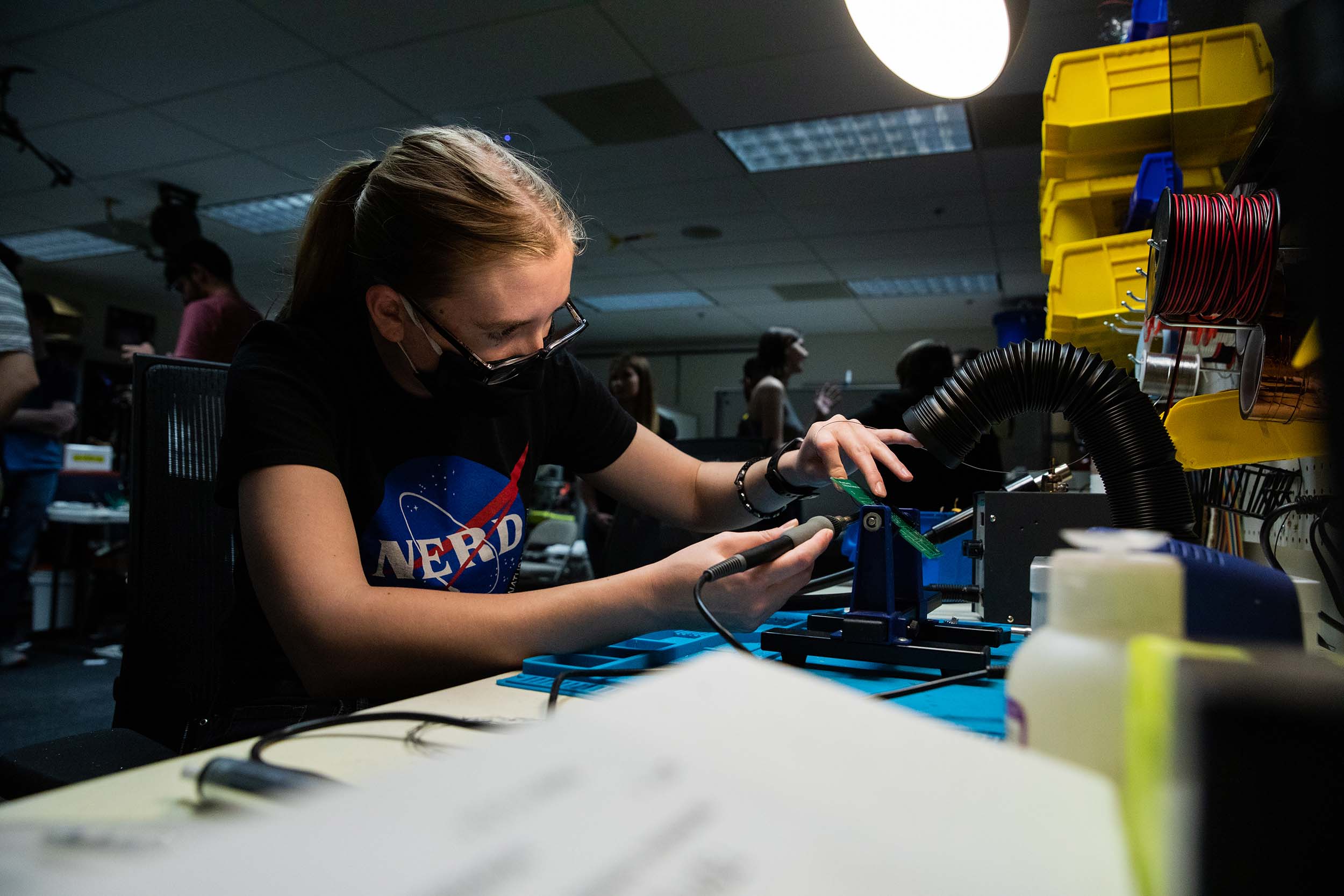 A female student wears a shirt bearing an altered "NASA" logo with "nerd" replacing "NASA," while soldering in the Luminosity Lab.