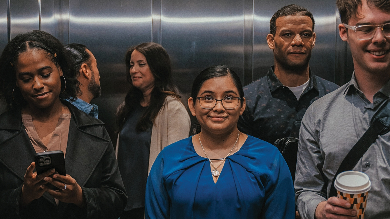 A diverse group of people in professional attire inside an elevator, with one person smiling at the camera while another looks at a smartphone.