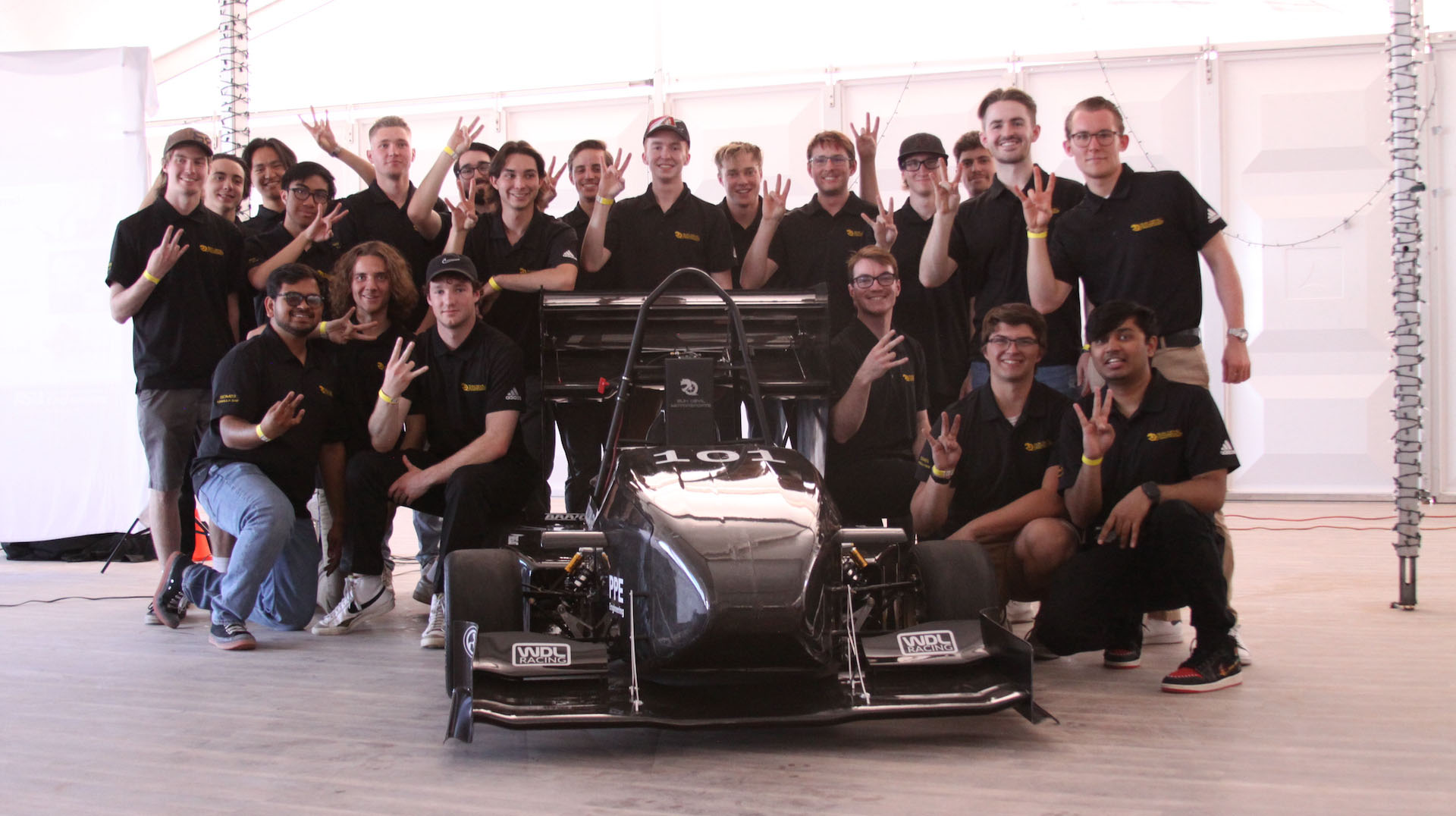 The members of the Sun Devil Motorsports team pose for a photo, displaying the ASU pitchfork school pride gesture, surrounding their latest race car, the SDM23.