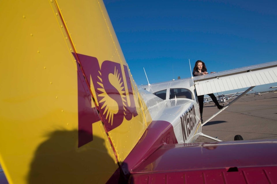 A female student stands on the wing support structure of a small ASU airplane, inspecting the top of the wing