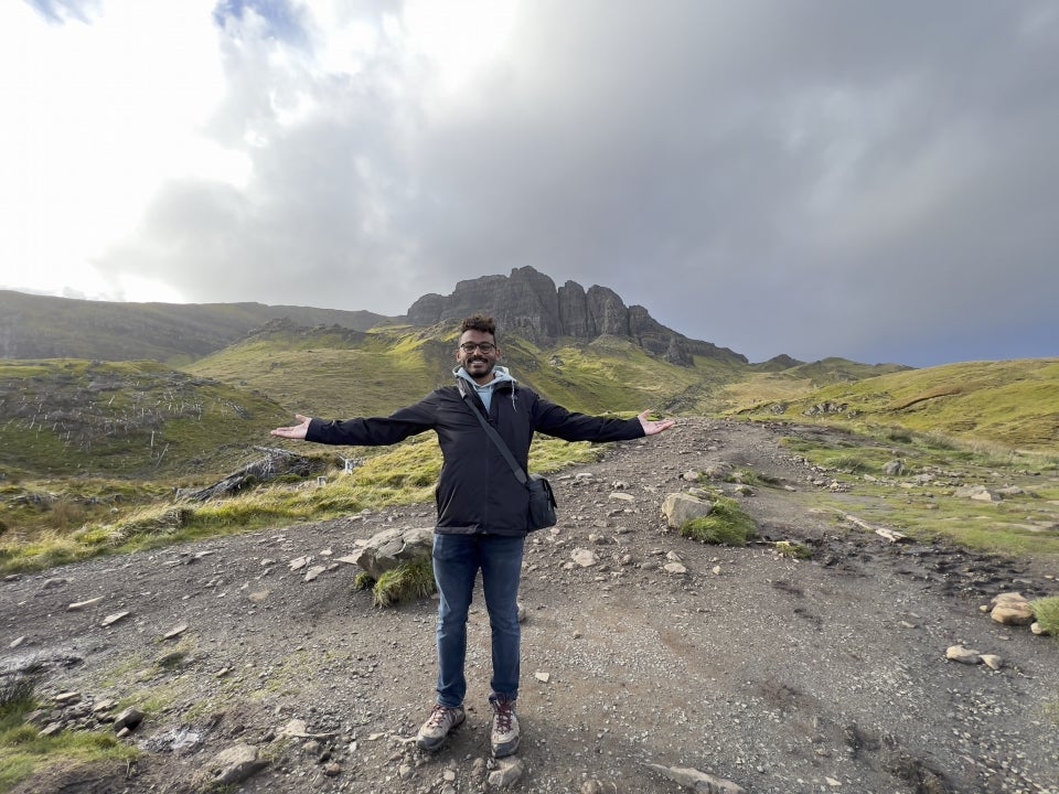 A male Fulton Schools student stands alone, arms wide the the sky and smiling, in a remote location with a mountainous terrain behind him