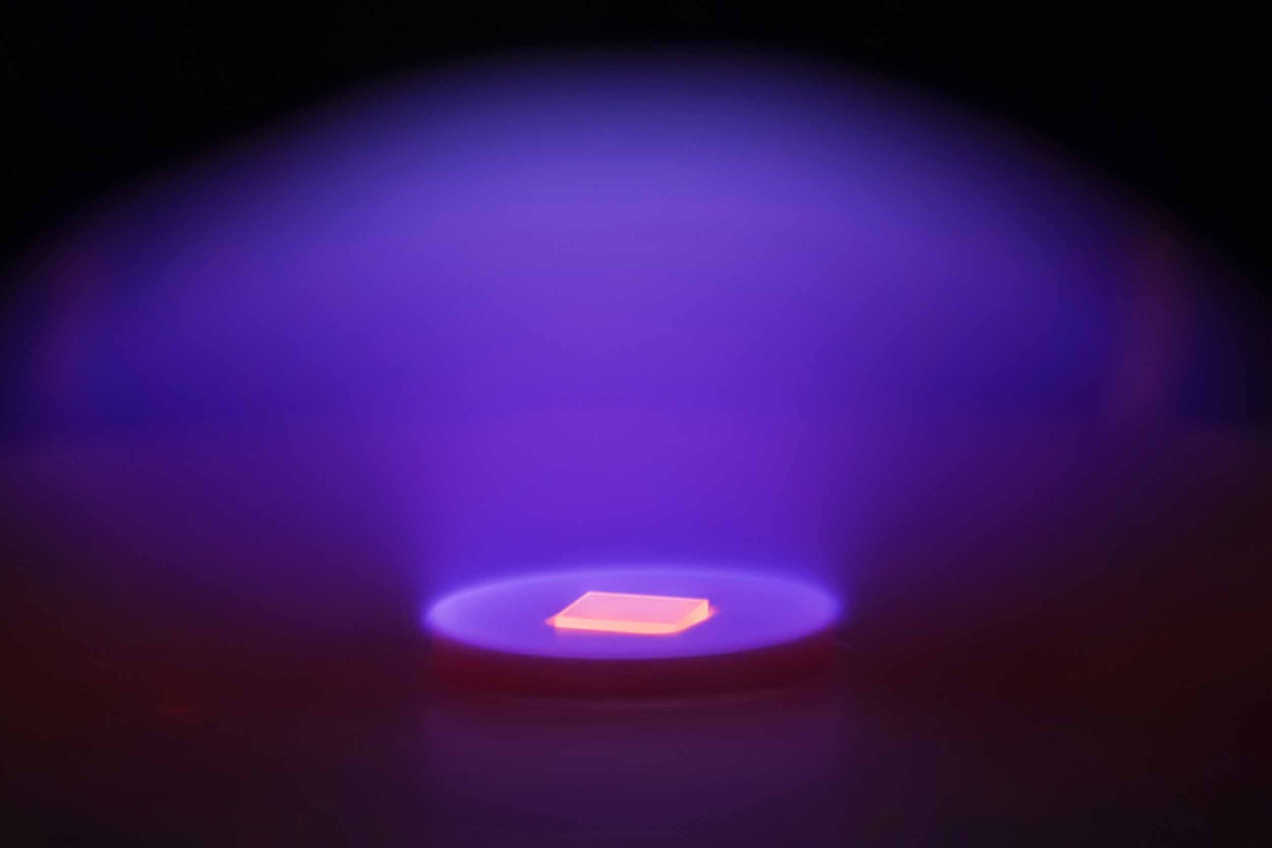 A microscopic photo of an artificial diamond growing in a lab environment. It looks like an golden, glowing, flat but layered square illuminated by a purple glowing disk.