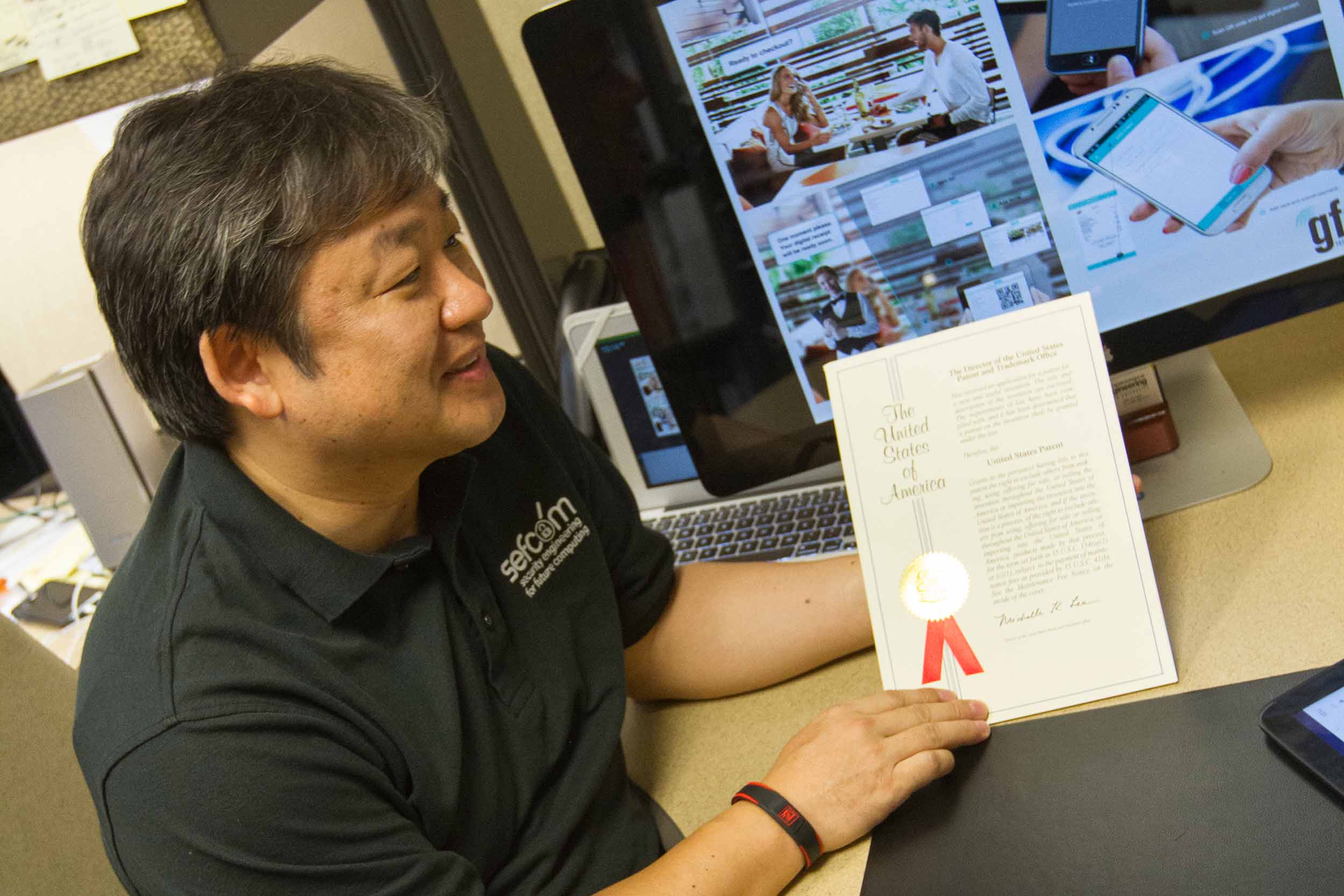 Gail-Joon Ahn sits at his desk, smiling, holding an award certificate in his hand.