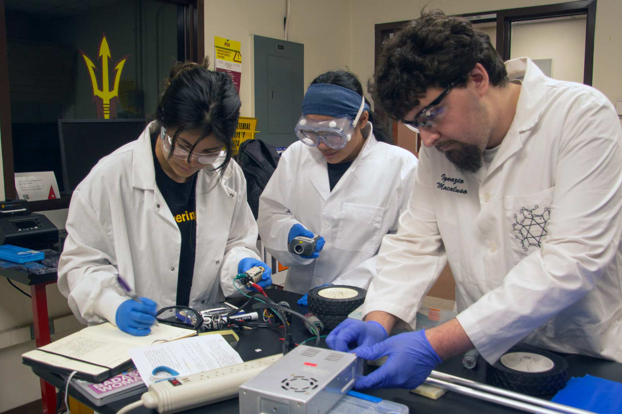 Three students in lab safety gear work at a bench on an electronic object.