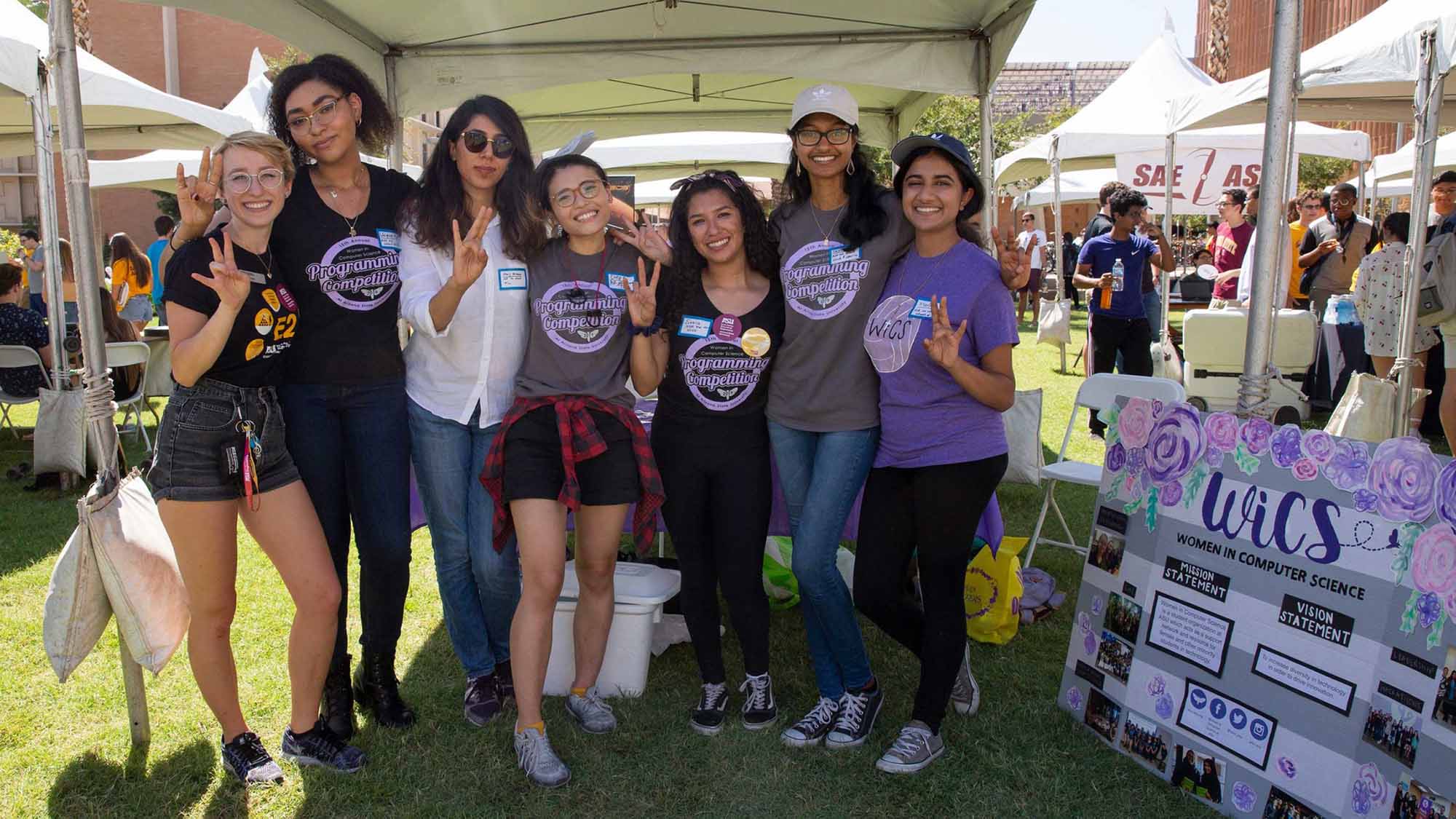 A group of Women In Computer Science club members stand together, arm in arm, at their booth at the Sun Devil Welcome event.