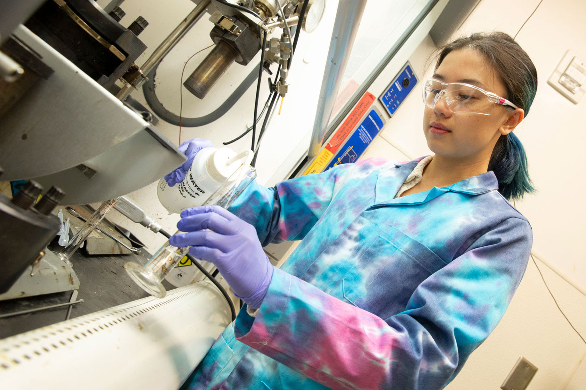 An ASU Engineering student researcher wears a tie-dyed lab coat, protective goggles and lavender latex gloves while working in her lab under the ventilation hood.