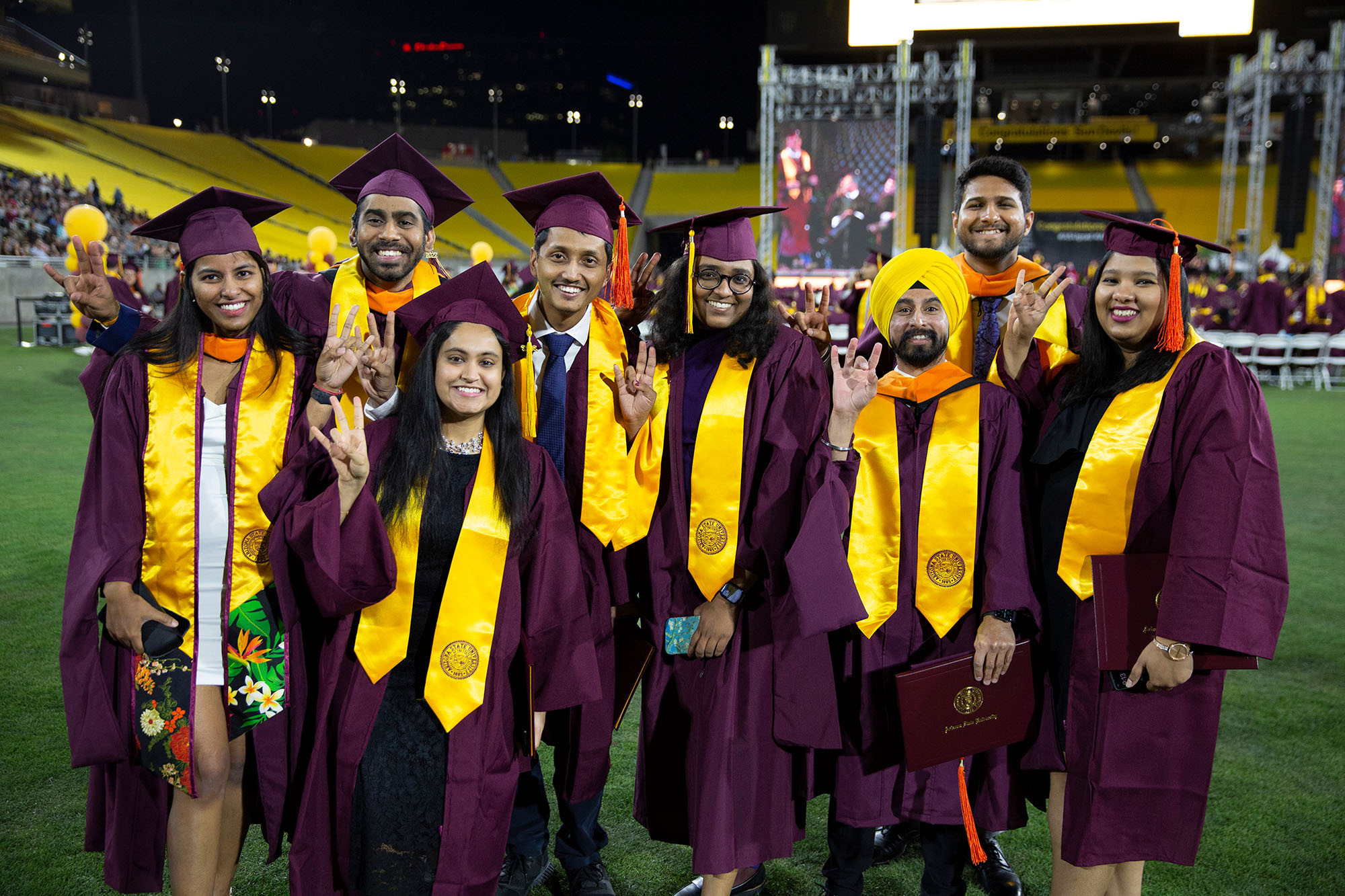 A group of 8 happy grads stand together for a group photo, each showing the ASU pitchfork school spirit gesture and smiling big for the camera on the field at Sun Devil Stadium.