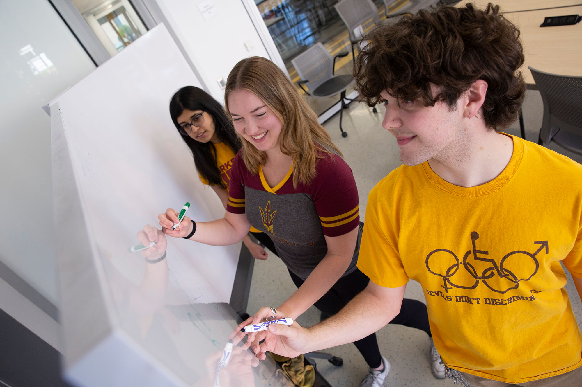 Three ASU students, two women and one man, stand at a white board, writing.