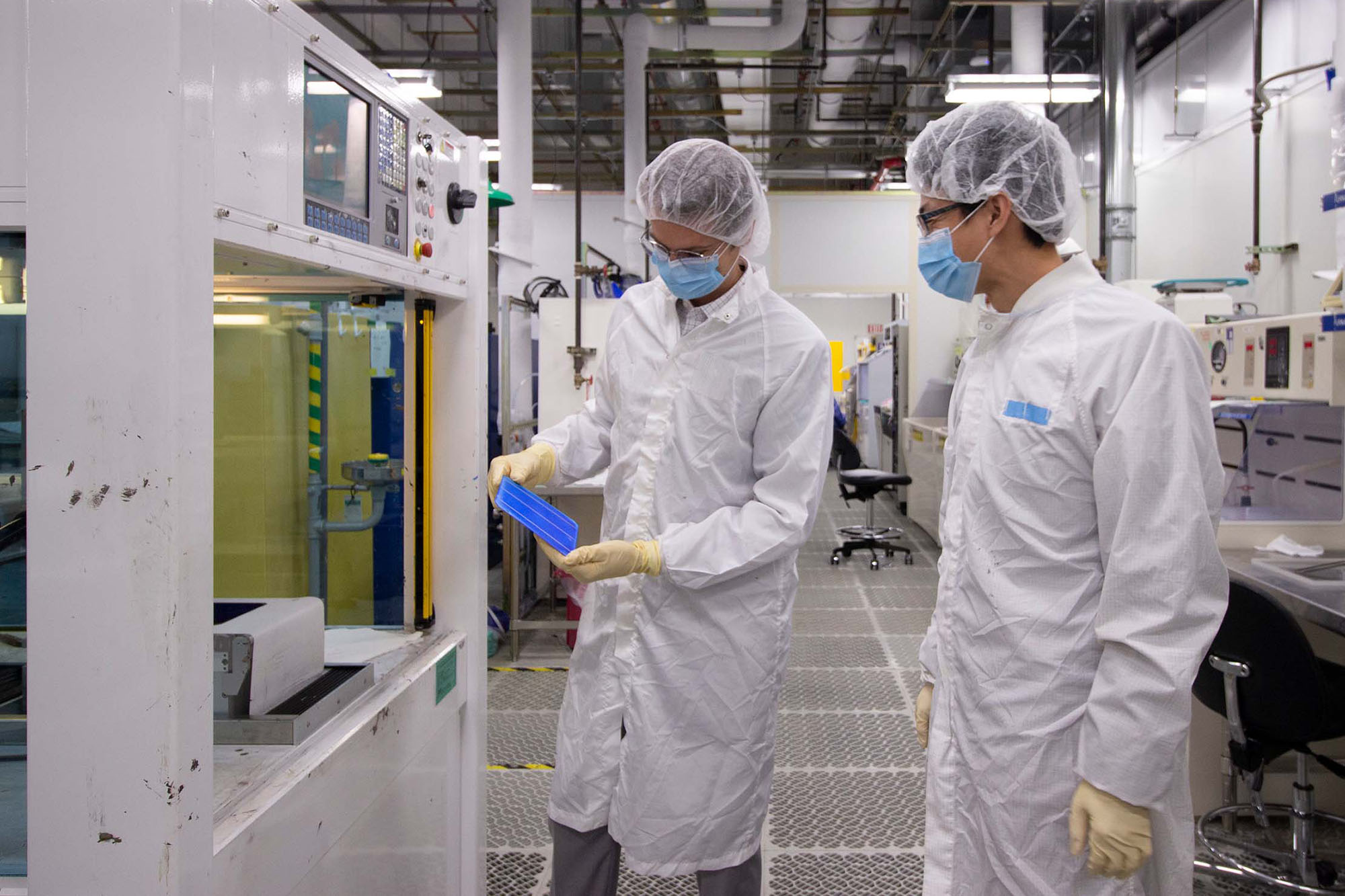 Two researchers wearing protective clothing (hairnets, masks, gloves and lab coats) examine an object one is holding inside of a very large research facility.