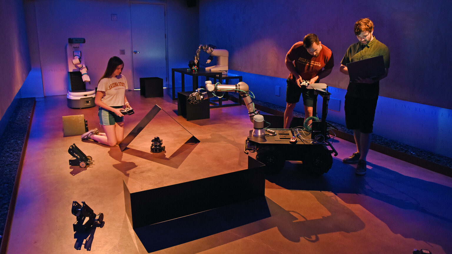 Three ASU students work in a mood-lit room with several robots, having them perform tasks under different conditions.