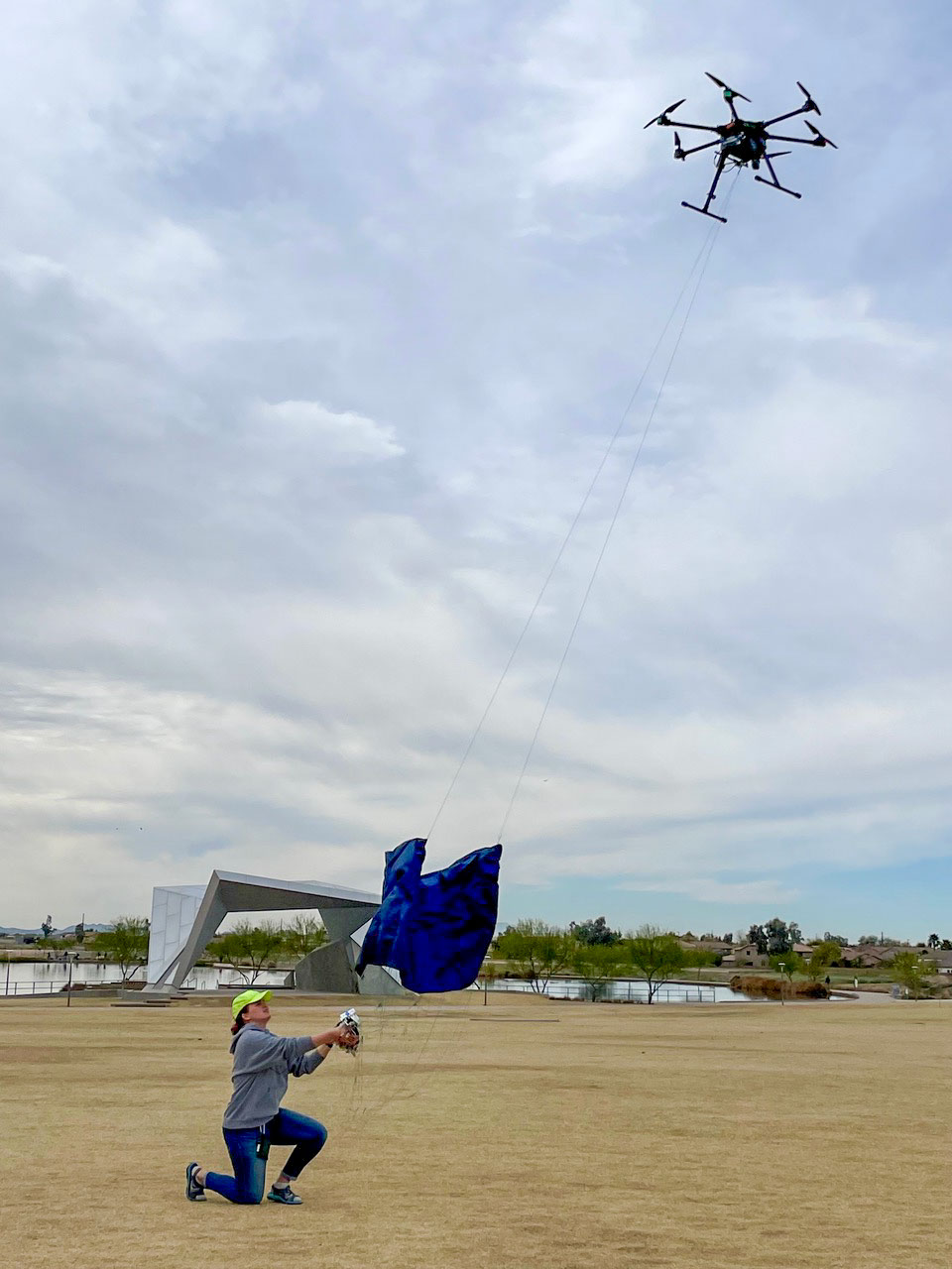 A woman on the ground interacts with a large drone in the sky, testing a drone-driven parachute deployment system.