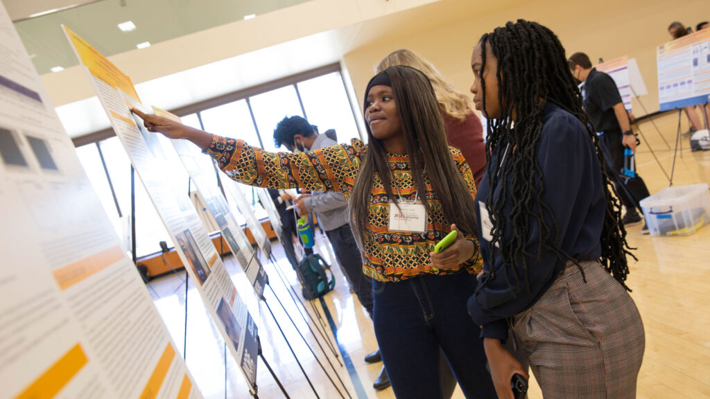 Two student researchers stand at their research poster during a research expo.