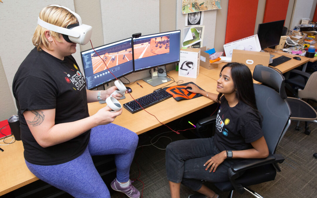 Two students sit together at a computer terminal, with one of them wearing a pair of VR goggles.