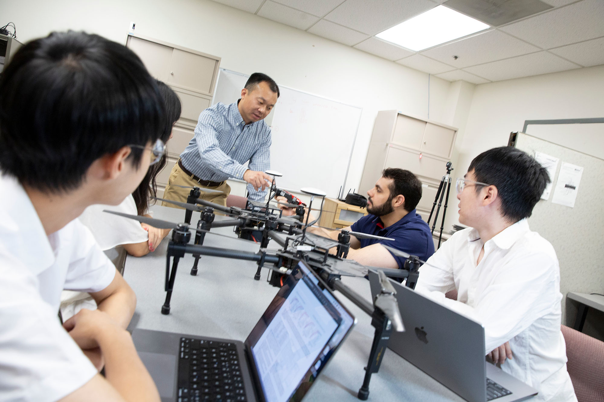 A professor talks to a group of students assembled around a large rectangle table with drones on the table.