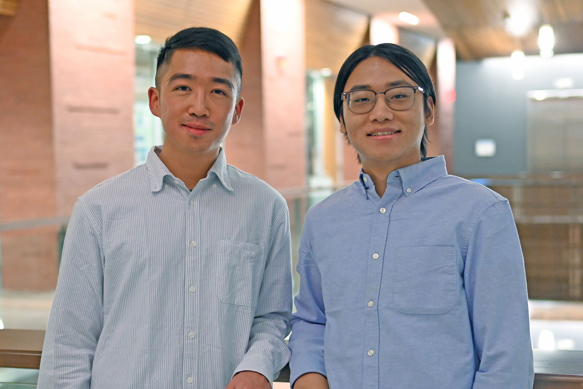 Two men dressed in two shades of light blue Oxford shirt, stand side by side, slightly smiling for the photographer.