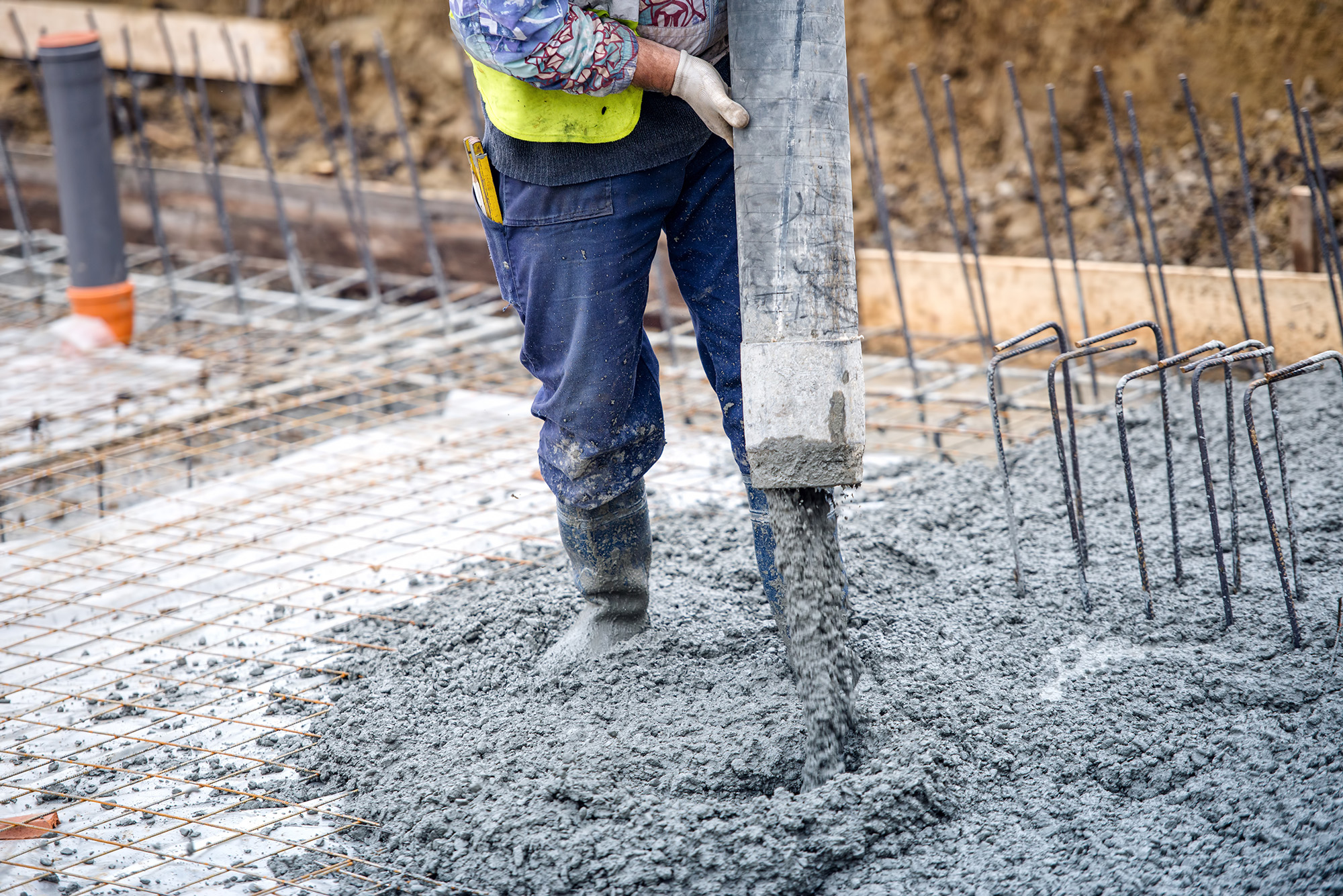 A person directs a large tube coming off of a concrete mixer that is pouring concrete into a prepared area on the ground.