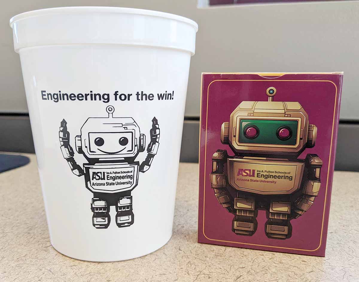 A white Fulton Schools branded stadium cup sits next to a maroon pack of cards with a robot displaying the Fulton Schools logo on it. 