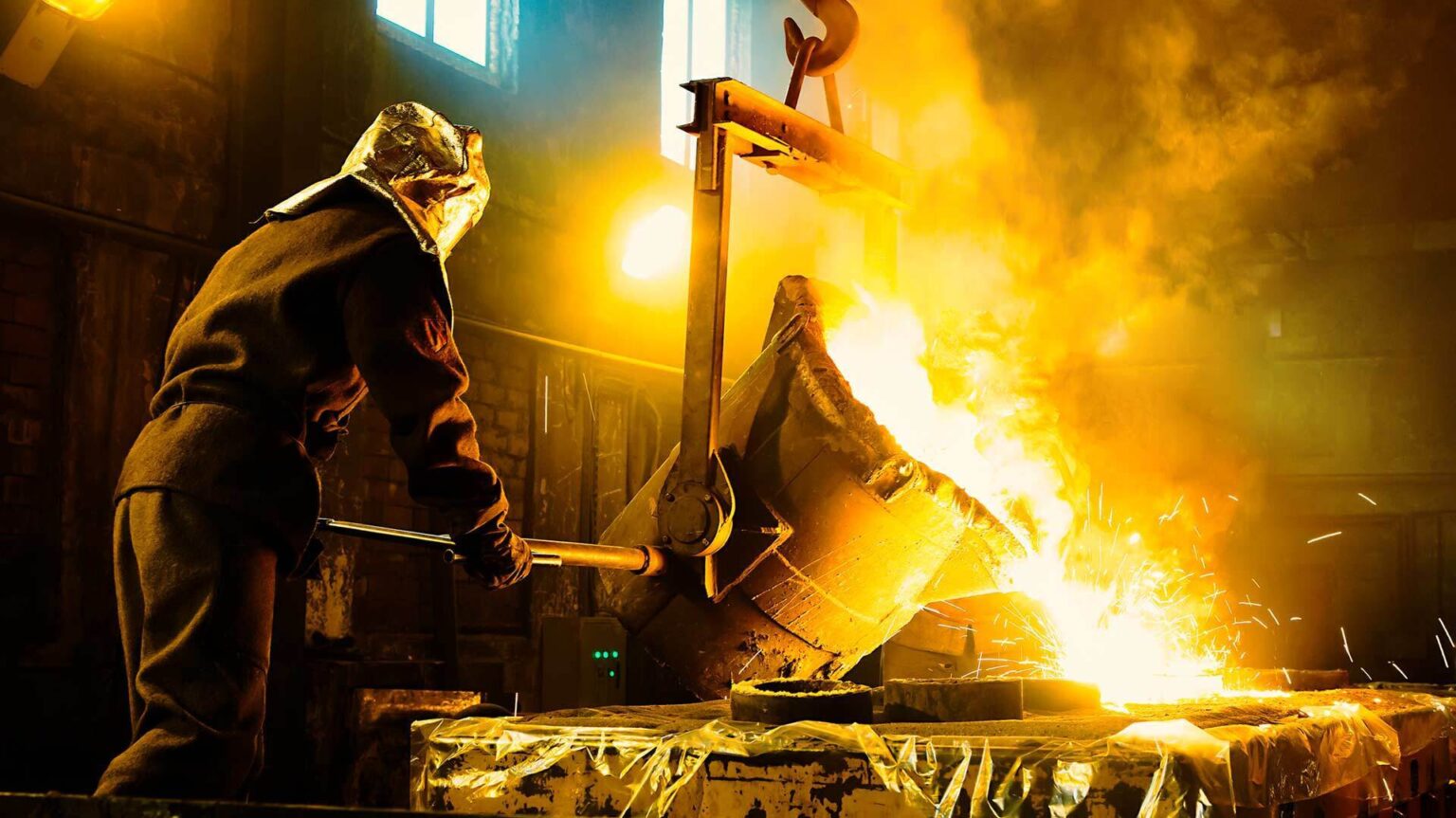 A steel worker wearing protective clothing guides the pouring of a vat of molten metal inot a container.