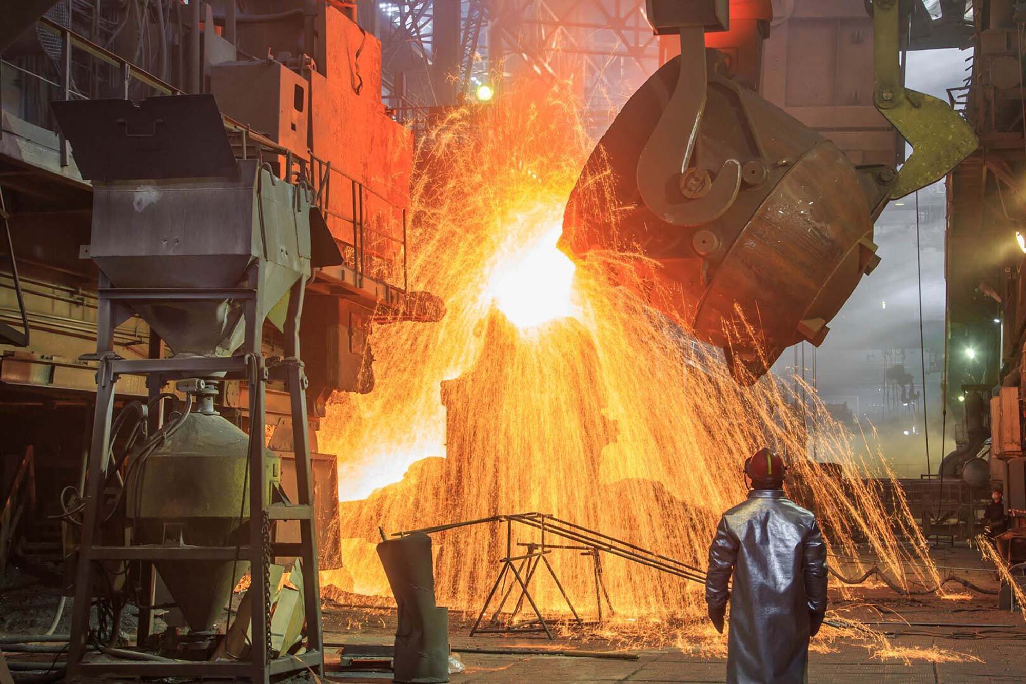 Stock image of a large vat of molten metal being poured into some kind of mold. Sparks are flying everwhere and a man in a reflective protective clothing is looking on