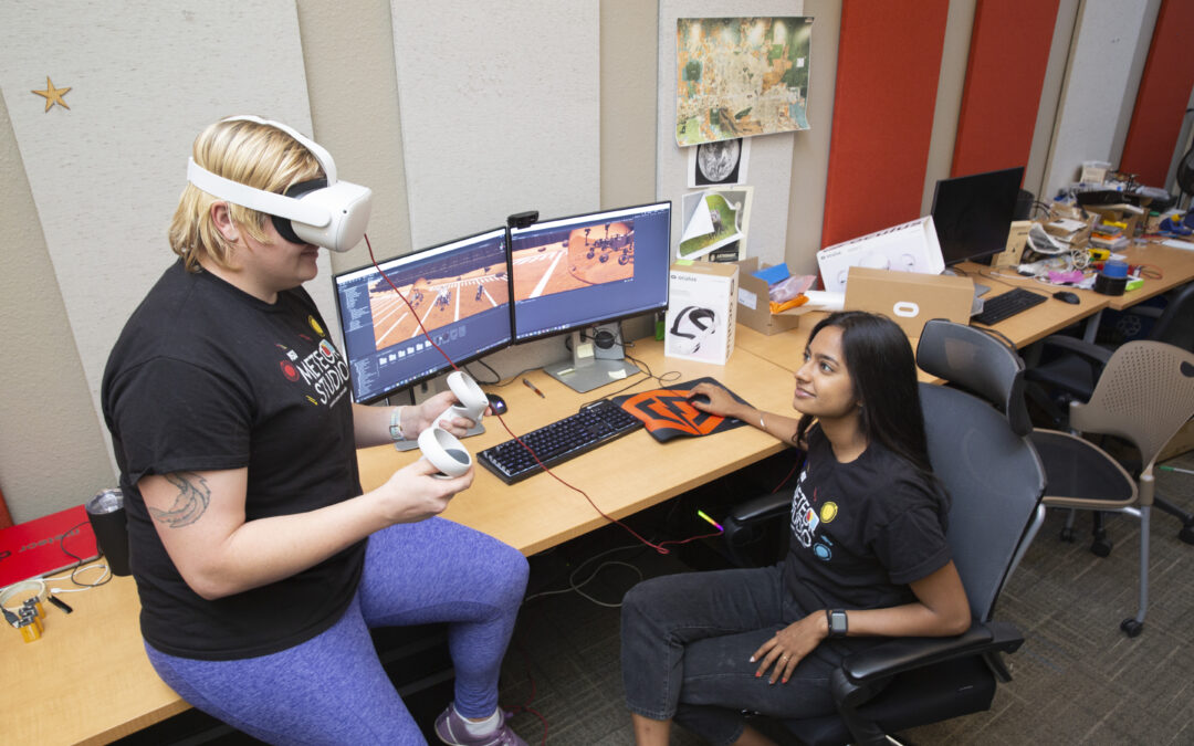 A student uses VR glasses while another monitors a computer.