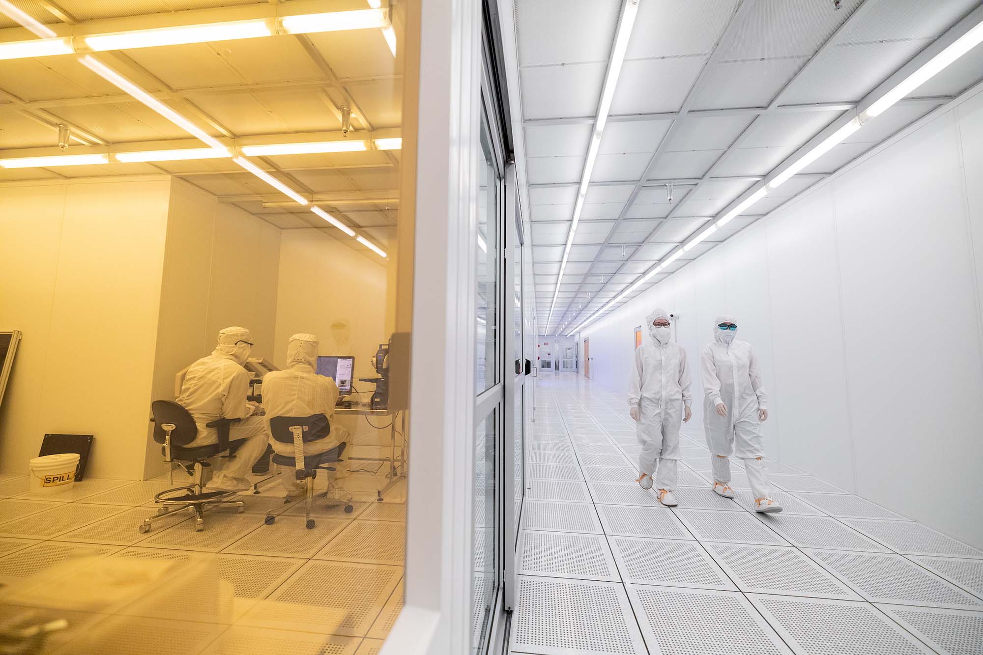 A view of a hallway (right) inside ASU's Macrotechnology Works research facility. On the left is a view into a room where the lighting is yellow. On both the right and the left, people are wearing clean room protective clothing.