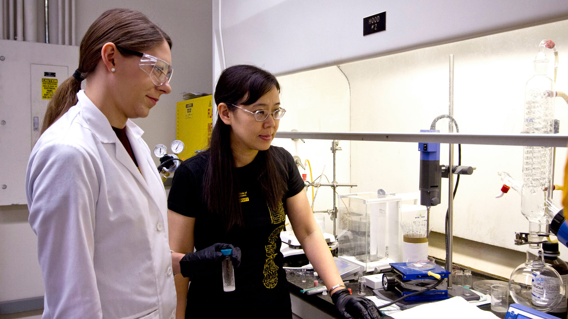 Two women stand in a research lab. One, on the right, advises the one on the left, a doctoral student wearing protective lab gear.