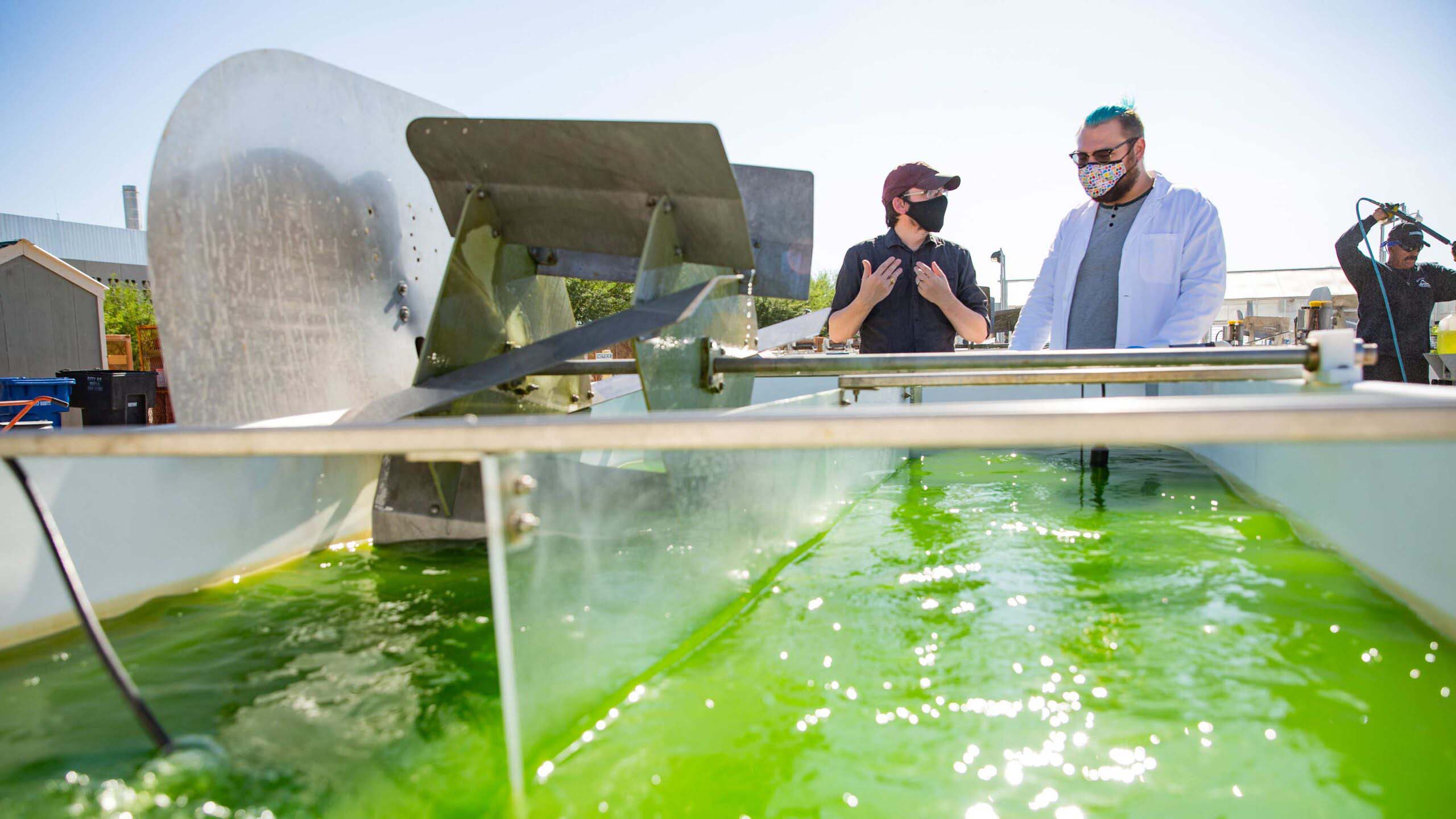 Taylor Weiss and Duane Barbano stand outside in the bright Arizona sun next to a gigantic vat of churning water full of bright green algae